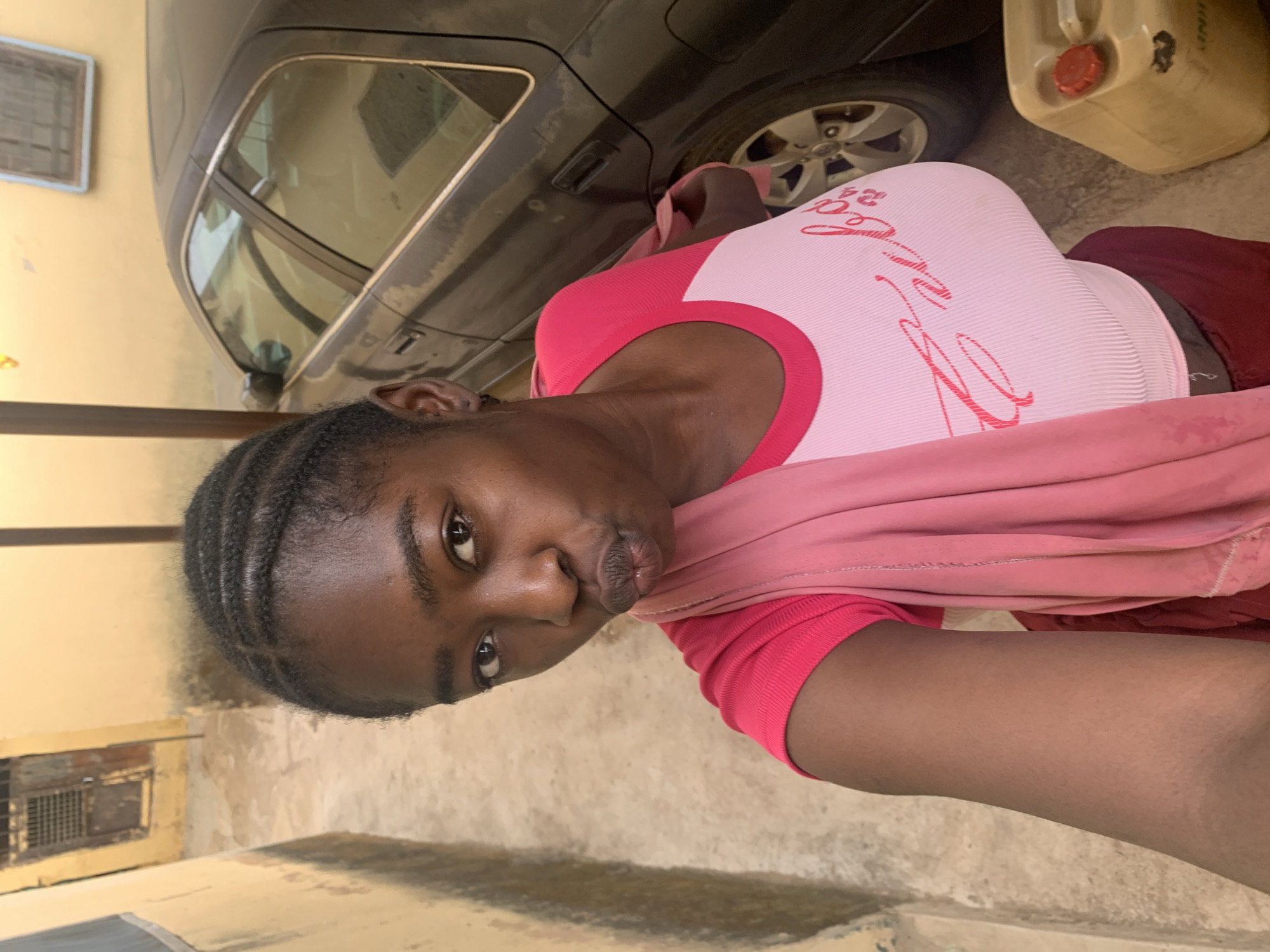 A young Black woman is holding a smartphone in a selfie pose, likely taken in Rafin Par, Nigeria. She has braided hair and is wearing a pink and white top with a pink shawl. The environment appears to be an outdoor area, with a car parked in the background. There is also a yellow gas container next to the car. The ground is paved and there is a building visible in the background with a window air conditioner and a drainage vent.