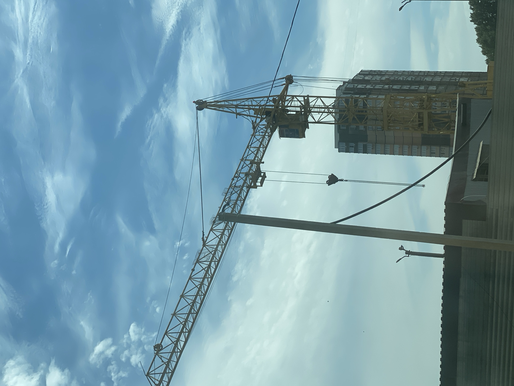 A yellow construction crane is prominently featured in the foreground, its long boom extending diagonally across the frame against a bright blue sky with scattered white clouds. Below the crane, a tall, multi-story building with a dark facade and many windows is under construction. In the lower part of the image, a corrugated metal structure and a partial view of a fence are visible. The scene suggests an active construction site within an urban environment, possibly on a clear day.