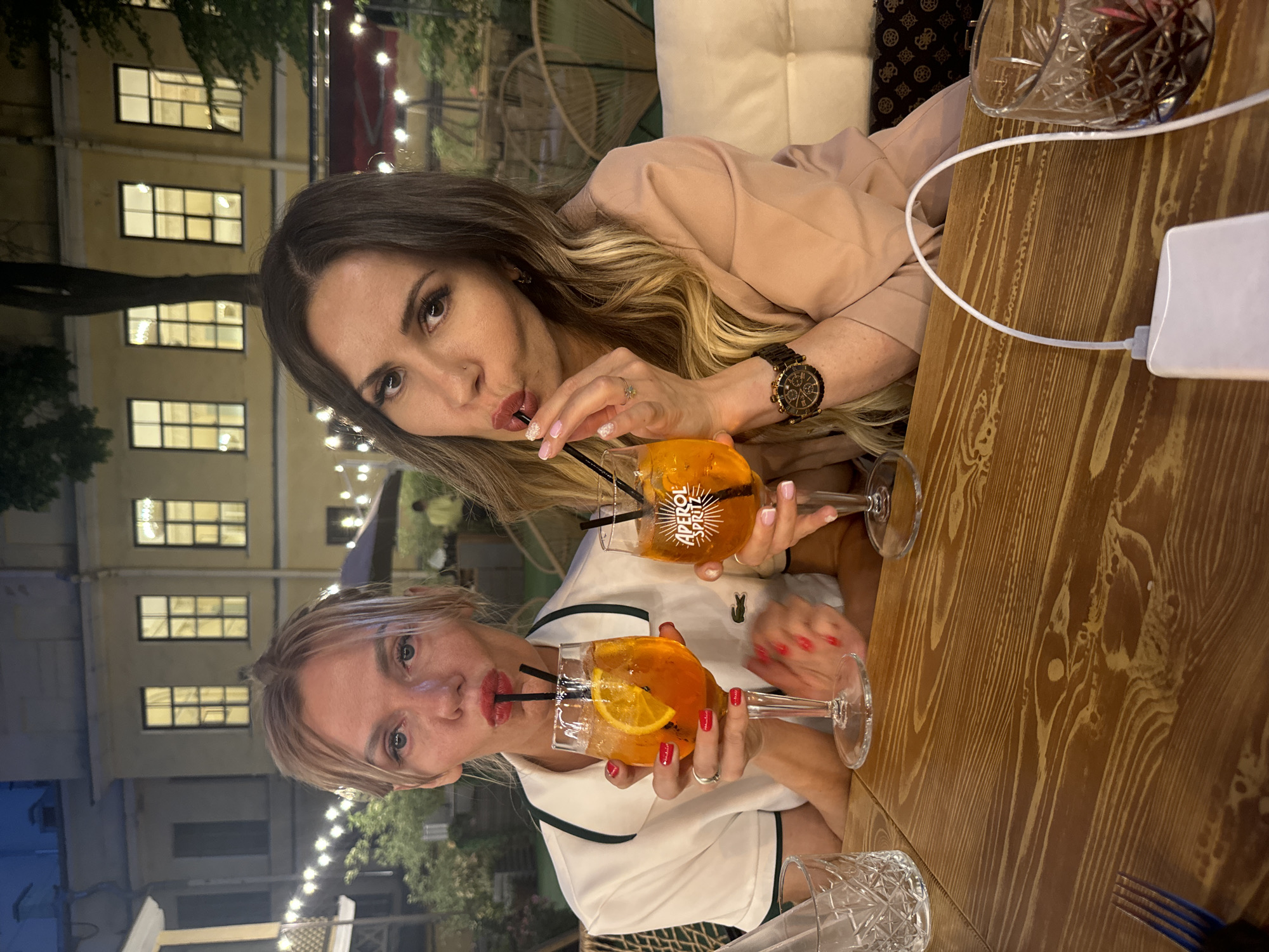 The image captures two adult females seated at a wooden table in an outdoor restaurant or cafe setting during the evening or night, located in Saint Petersburg, Russia. Both individuals are looking directly at the camera, posing with pursed lips around black drinking straws that extend into their respective orange cocktails.
The woman on the left has light-colored hair, pulled back, and is wearing a white short-sleeved polo shirt with dark green trim and a small crocodile logo on her chest. She holds an Aperol Spritz cocktail, garnished with an orange slice, in a stemmed glass. Her fingernails are painted red, and a silver ring is visible on her left ring finger.
The woman on the right has long, wavy dark blonde or light brown hair with highlights. She is dressed in a light peach or beige blazer. She holds a stemmed glass prominently displaying "APEROL SPRITZ" on its side, also containing an orange cocktail. A black and gold chronograph watch is visible on her left wrist, and a small stud piercing is present on her left nostril. Her fingernails are painted a light color, and a small ring is on her right ring finger.
On the wooden table, a clear, patterned glass tumbler and a blue fork are partially visible on the left. On the right, a white power bank with a white USB cable is situated next to another patterned glass tumbler.
The background features a multi-story, light yellow building with several brightly lit rectangular windows, suggesting classical architecture. Numerous string lights illuminate the outdoor area, revealing green foliage, dark umbrellas, and various outdoor seating options, including wicker-style chairs. A segment of a red awning or similar structure is visible in the upper right background.