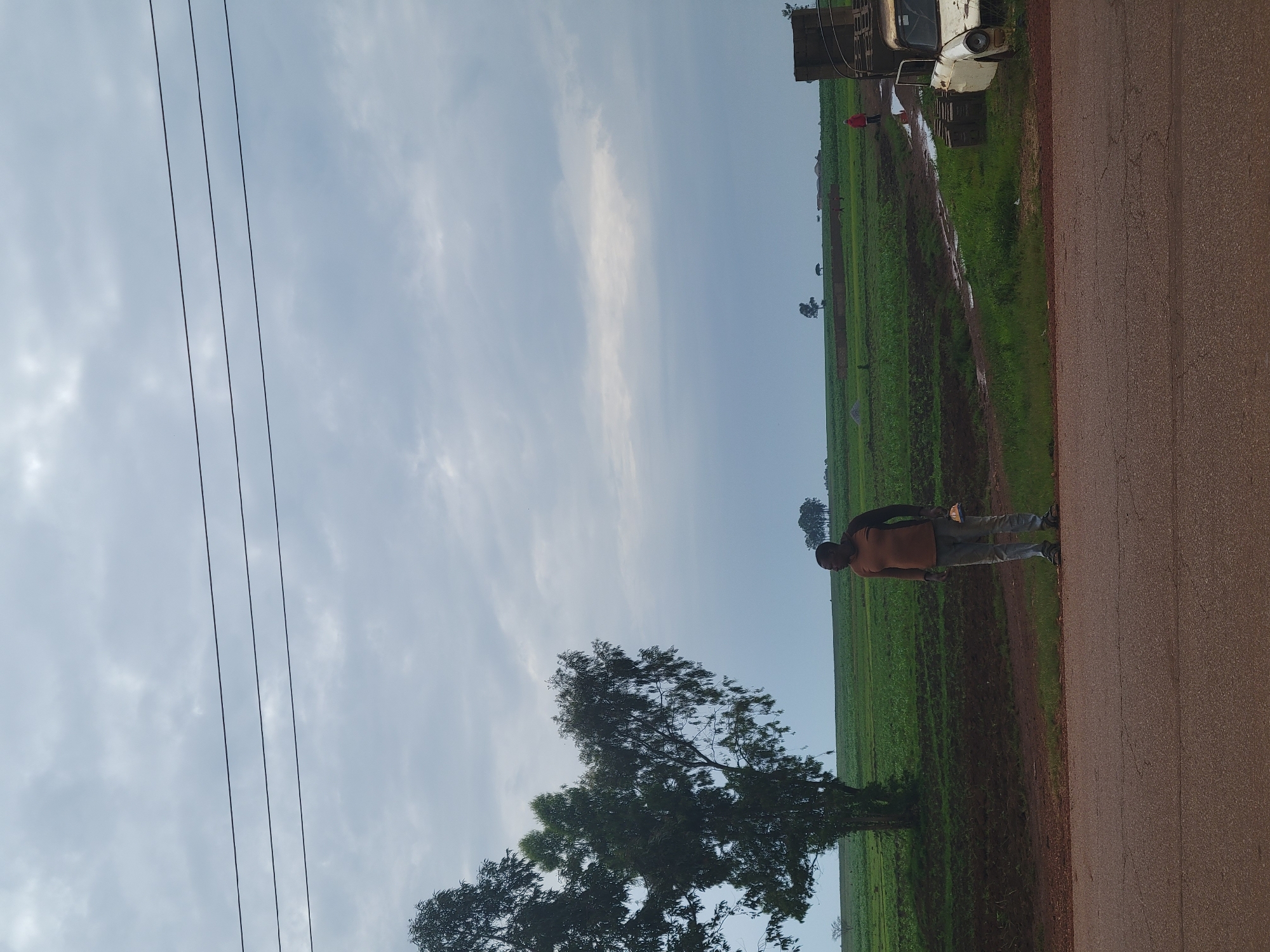 A man stands on a dirt road in front of a vast green field under a cloudy sky. The man is wearing a brown shirt and grey pants, and he is holding a white object in his right hand. To the right of the man, a rusty white car with its door open is parked near a stack of bricks and wooden pallets. The field is a lush green, with rows of crops visible. In the distance, several trees and a few small buildings are scattered across the landscape. Three parallel power lines stretch across the upper portion of the image. The sky is overcast with a mix of white and grey clouds.
