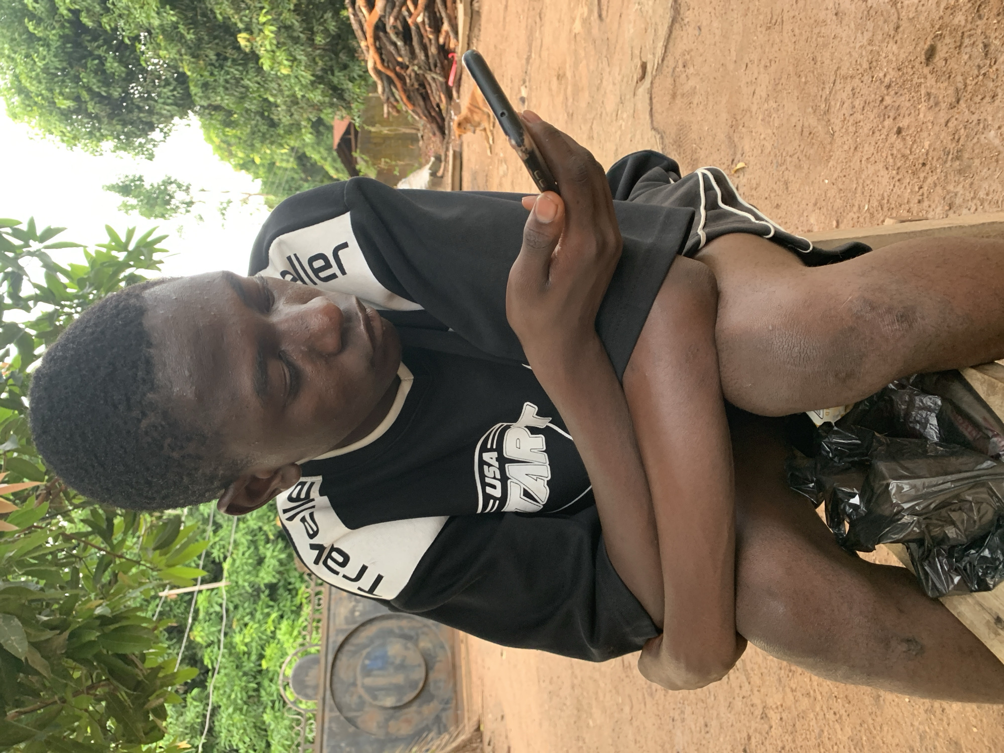 A young man is seated outdoors, likely in a rural area on the outskirts of Akwanga, Nigeria, based on the environment and the subjects ethnicity. He is looking at his phone, while sitting on a wooden structure of some sort. The background shows trees and a dirt area, with a pile of cut branches or sticks. The sky is partially visible through the trees, suggesting it may be daytime. A black trash bag is seen on the edge of the wooden structure he is sitting on. The scene suggests a simple, possibly impoverished setting.