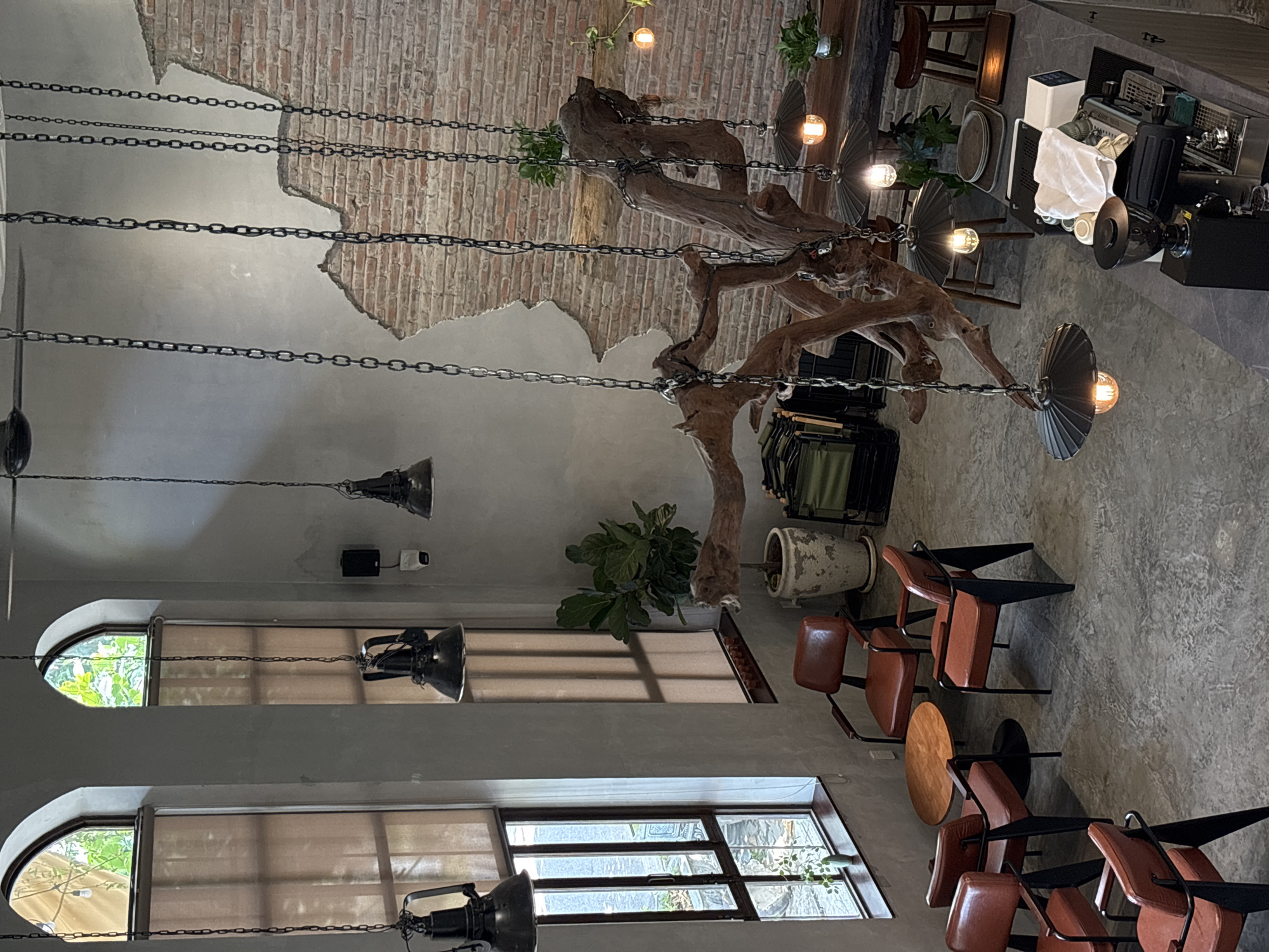 The image depicts the interior of a coffee shop in Da Nang, Vietnam. The space features a rustic and industrial aesthetic with exposed brick walls, concrete floors, and high ceilings. Large, arched windows with sheer blinds allow natural light to filter in. Hanging from the ceiling are several industrial-style pendant lamps with metal shades, suspended by chains.
The furniture includes several mid-century modern-inspired chairs with red leather upholstery and black metal frames, arranged around small, round wooden tables. A large, gnarled tree trunk, artfully displayed as a sculptural element, hangs from chains in the center of the room.
In the background, a bar area is visible, equipped with a coffee machine and other bar accouterments, suggesting it is an operational coffee shop. Plants are strategically placed throughout the space, adding a touch of greenery to the industrial decor. The overall impression is a stylish, contemporary cafe designed for a relaxed ambiance. There are no people visible in the image.