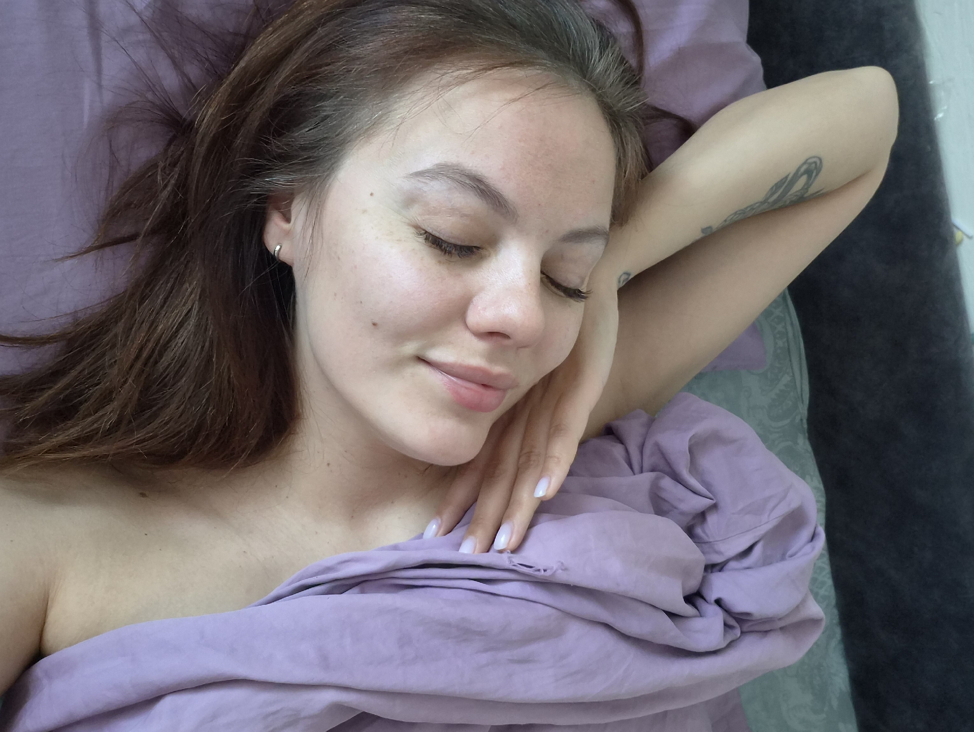 A woman is lying in bed. She has brown hair and closed eyes, with freckles visible on her face. She is wearing a small silver earring in her left ear. Her left arm is raised, with her hand resting near her chest. A tattoo is visible on her forearm. She is covered with a purple-colored sheet. The background appears to be part of a bedroom. The photo was taken in the city of Chaikovsky, Russia.