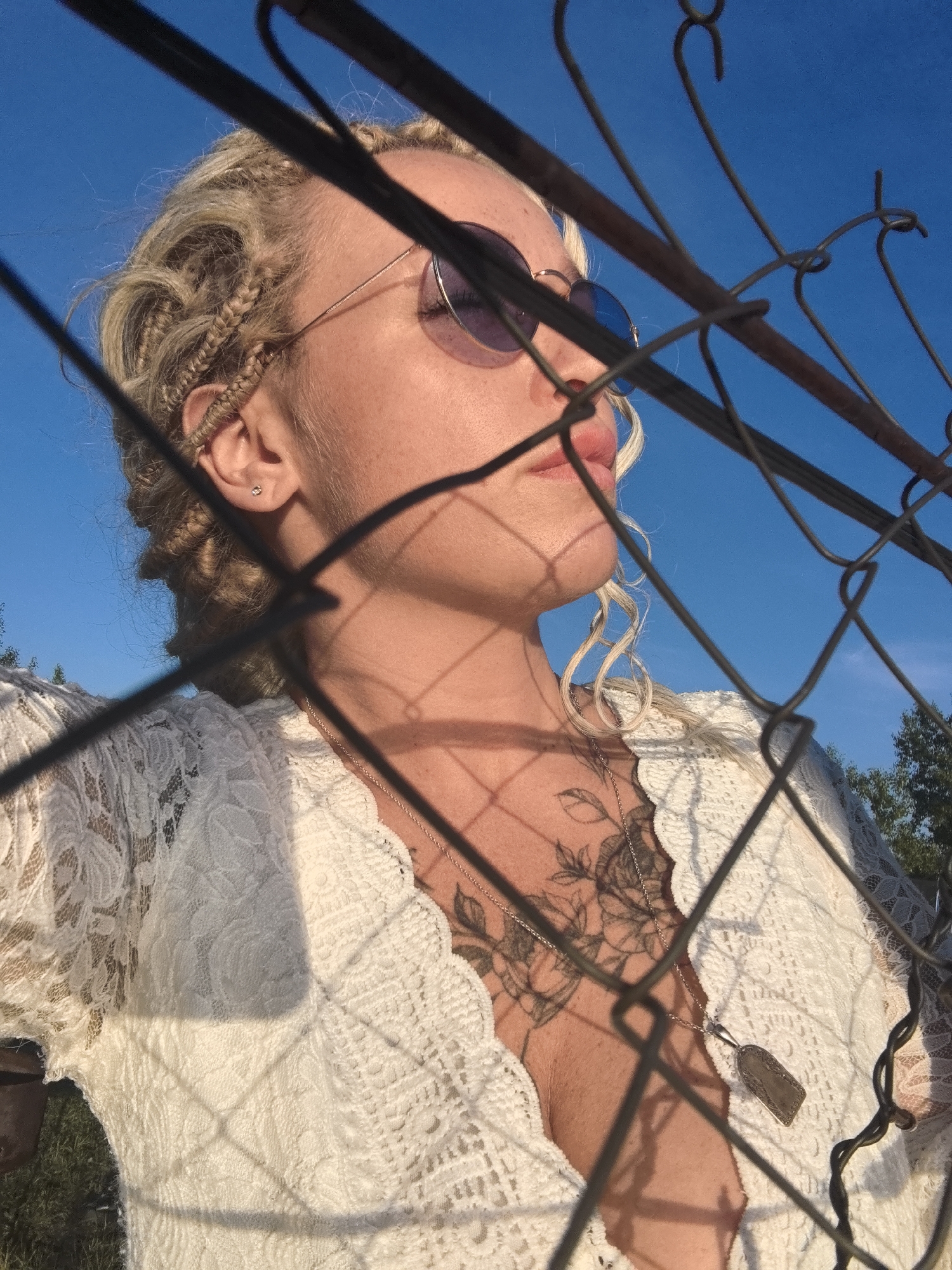 The image features a close-up profile of a young woman with blonde hair styled in braids and loose curls. She is wearing round, purple-tinted sunglasses and a white, lace top with intricate floral patterns. A silver necklace with a rectangular pendant rests on her chest, over a tattoo of roses. She has a small, stud earring in her visible ear. The scene is outdoors, with a clear, bright blue sky as the background. The lighting suggests it is late afternoon or early evening, with warm sunlight casting shadows on her face and clothing, as well as creating a grid-like shadow from a wire fence in the foreground that partially obscures her. There are glimpses of green foliage and trees in the distance. The woman's expression is serene as she looks slightly upwards and away from the camera, possibly enjoying the moment.
