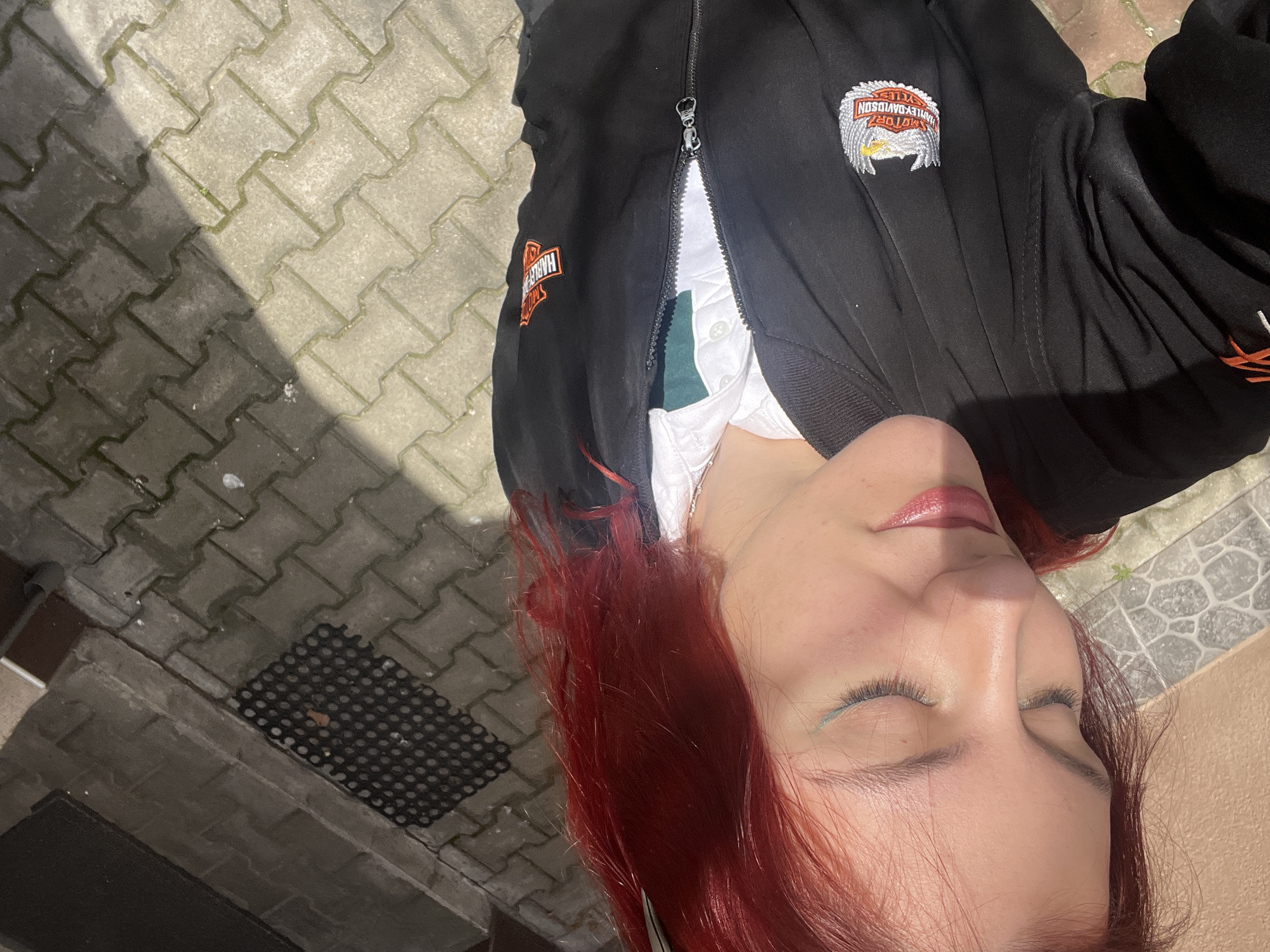 A young woman with red hair, wearing a black Harley Davidson jacket, is lying on a paved surface. Her eyes are closed, and she is wearing makeup. The paved surface appears to be a sidewalk or patio. A black rubber mat is nearby. The location is unknown and not verifiable with the given information. The city of Asenovgrad, Bulgaria, cannot be confirmed.