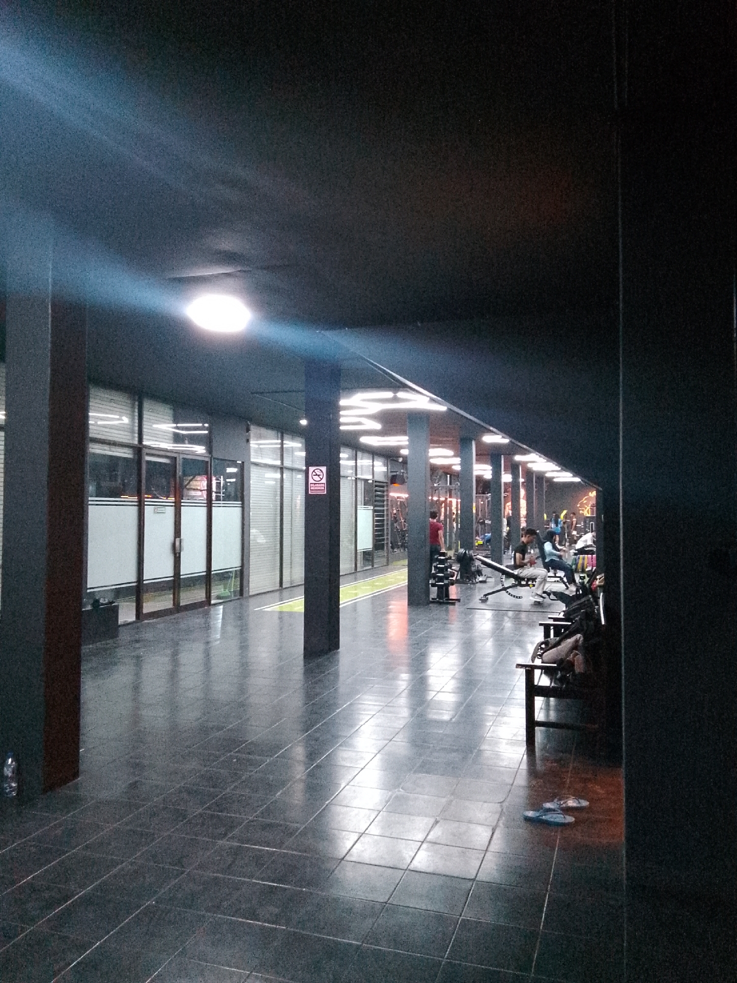 The image depicts the interior of a gym in Trenggalek, Indonesia, during the evening. The perspective is from a hallway leading into the gym area. The hallway floor is tiled with dark, reflective square tiles, creating a glossy surface. Overhead, dark ceiling panels are visible, interspersed with bright, circular fluorescent lights that cast a strong glow.
On the left side of the hallway, there are glass doors with frosted horizontal stripes and wooden frames, leading into what appears to be a reception or office area. A dark trash receptacle is placed near the entrance.
The hallway opens into the gym itself, which is equipped with various exercise machines. Several individuals are present, engaged in different activities. In the mid-ground, on the right side of the gym area, a man sits on a weight bench, looking towards the right. To his left, a person is seated on a similar bench, facing away from the camera. Further back, other gym-goers are visible, some appearing to be using machines or resting.
The gym's flooring is also tiled, with some areas marked with green lines. The lighting in the gym is a combination of bright overhead lights and the ambient light from the machines and possibly decorative elements.
In the foreground of the hallway, on the right, a section of a dark structure is visible, partially obscuring the view of the gym. On the tiled floor of the hallway, a pair of blue flip-flops are placed. The overall atmosphere is dim, with strong contrasts between the illuminated areas and the shadows cast by the ceiling and structural elements.