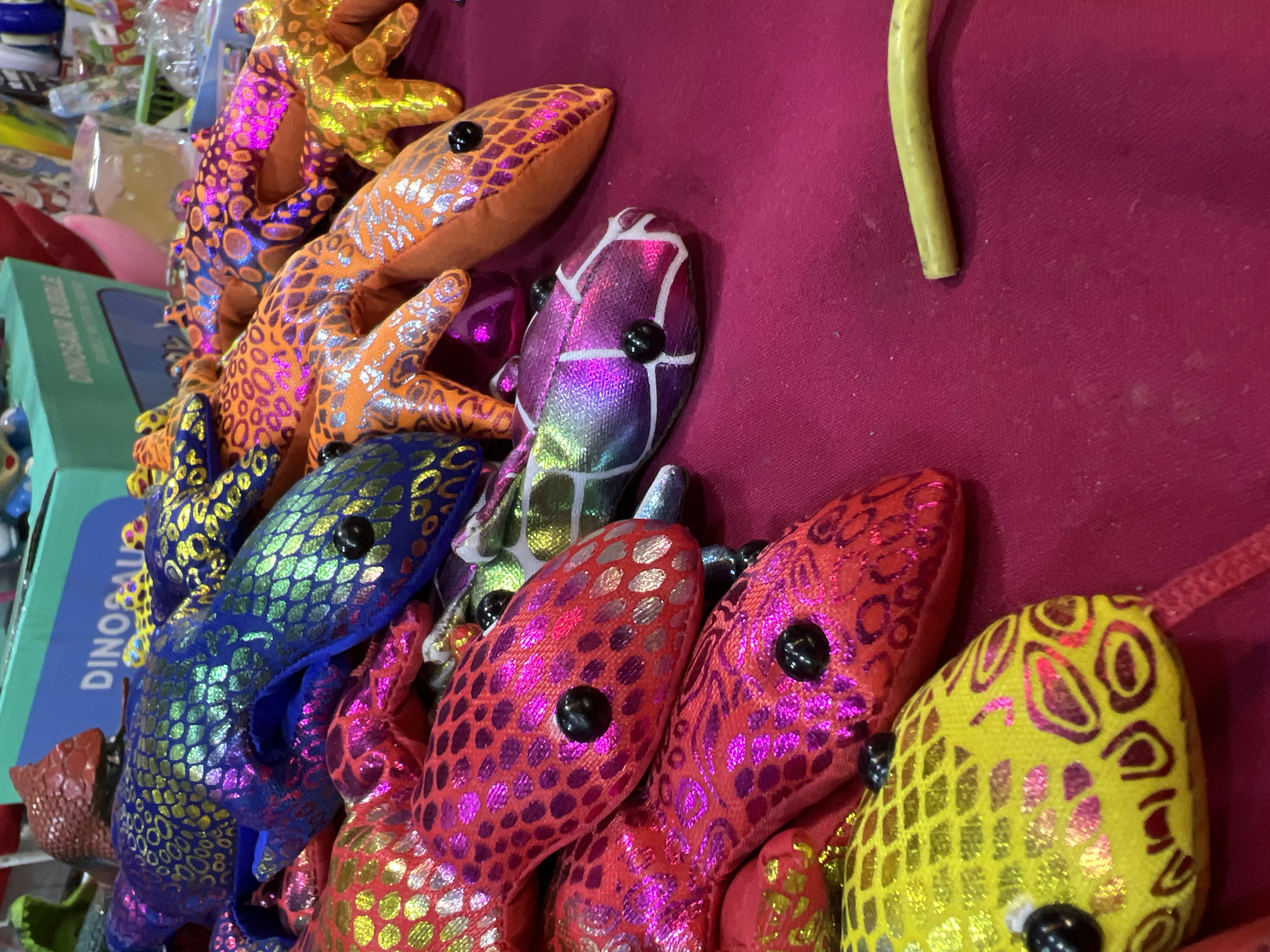 The image displays a collection of colorful, plush toy lizards with a glittery, iridescent finish. The lizards are predominantly blue, orange, pink, red, and yellow, each featuring black button eyes and a pattern of metallic spots or scales. They are arranged on a deep pink, textured surface, likely a tablecloth or fabric display. In the background, a green box with the word "DINOSAUR" is partially visible, suggesting a toy store or market setting. A yellow, tube-like object rests on the pink surface in the lower right portion of the frame. The scene appears to be an indoor display of merchandise.