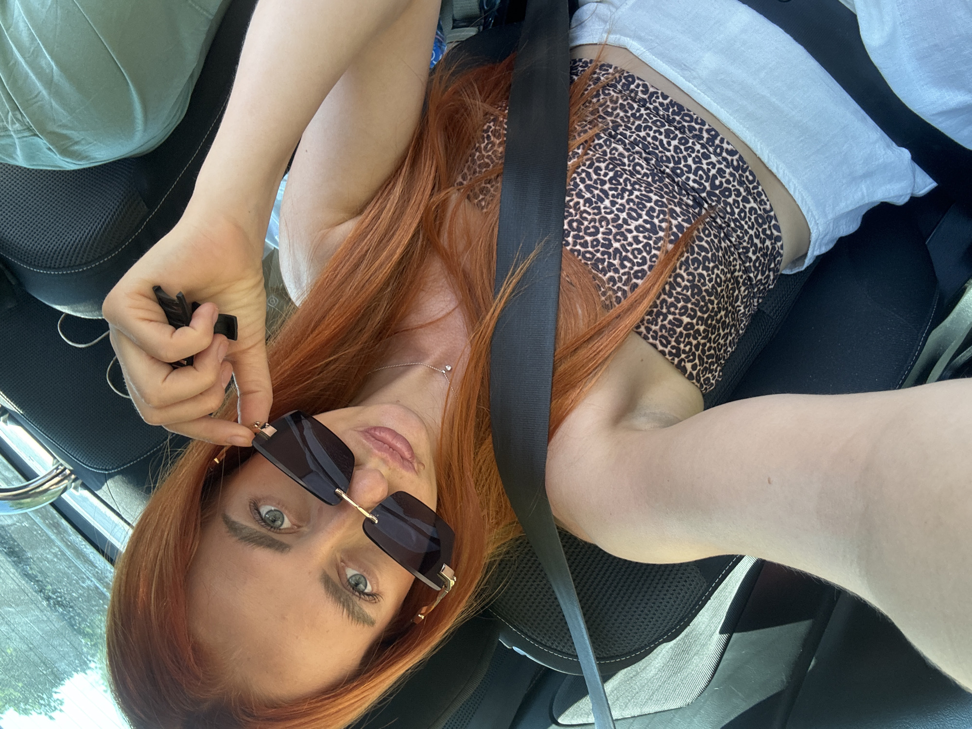 A woman with long, auburn hair is lying down in a car, her head tilted back and resting on the seat. She is wearing sunglasses with dark lenses and a gold frame, which she is holding up to adjust. Her top is a white tank top layered over a leopard-print bandeau. A black seatbelt crosses over her chest. Her eyes are light green and wide open, and her lips are slightly parted. She is wearing a delicate silver necklace with a small pendant. Her left hand is holding a small, black object, possibly car keys. The car appears to be parked, with the passenger seat visible and a glimpse of a window showing greenery and sunlight, suggesting it is daytime and the weather is clear. The scene conveys a relaxed, possibly candid moment.