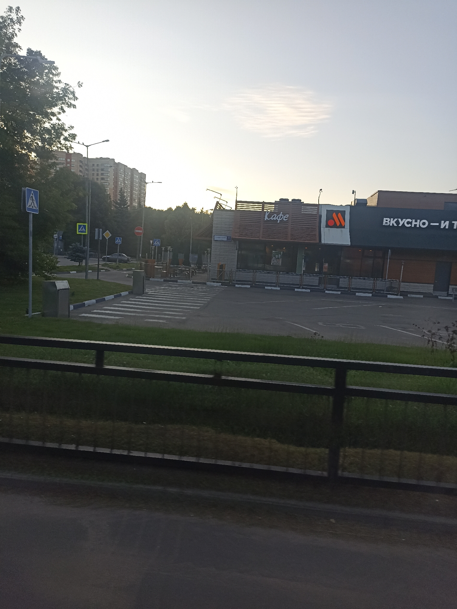 This is a photograph of a street scene in Chekhov, Russia, featuring a McDonald's restaurant and residential buildings. The McDonald's building has a prominent orange "M" logo and text in Russian that reads "ВКУСНО - И Т" (Vkusno - I T), which translates to "Tasty - And T". Adjacent to the McDonald's is a building labeled "Кафе" (Cafe). In the foreground, there is a paved area with pedestrian crossings, a grassy verge, and a black metal fence. A trash receptacle is visible to the left of the pedestrian crossing. In the background, tall residential buildings are visible, suggesting an urban or suburban environment. The sky is bright with some wispy clouds, indicating daylight. Traffic signs are present, including a pedestrian crossing sign and a no entry sign. A car is parked on the road.