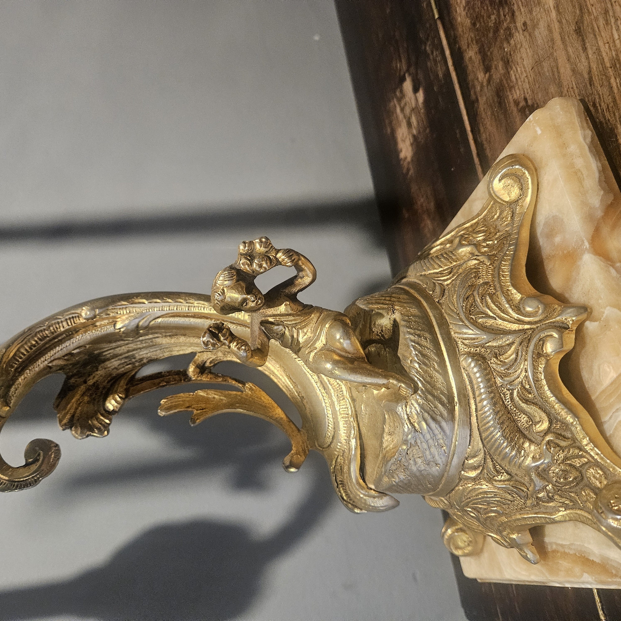 The image shows a close-up of an ornate, golden cherub figurine resting on a marble base, positioned on a rustic wooden surface. The figurine is made of metal, possibly brass or bronze, and features intricate detailing. A cherub with its arms raised and holding what appears to be flowers is depicted in the center of the sculpture. The cherub is sitting on a rough, rock-like texture that is part of the base. The base itself is decorated with scrolling acanthus leaves and other decorative motifs. The marble base is translucent with swirling patterns of beige and white. In the background, a grey wall with a shadow cast from the object is visible. The lighting suggests an indoor setting. The scene is not set in Almadrones, Spain, nor does it contain any elements that would suggest a specific urban location.