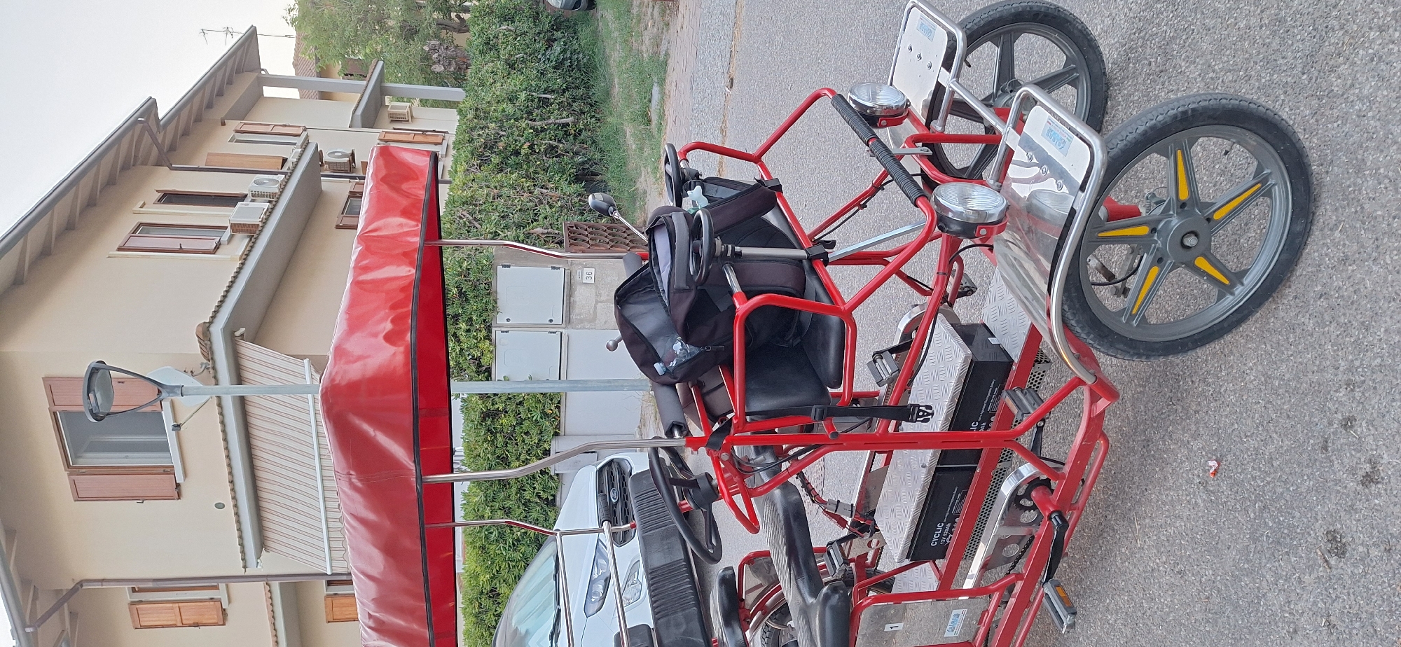 An electric quadricycle with a red canopy is parked on a paved surface in front of a multi-story building. The quadricycle has four wheels, two seats, and a steering wheel. A black backpack is placed on the passenger seat. The building appears to be a residential property with a light-colored facade, brown shutters, and air conditioning units. A hedge runs along the front of the property, and a street lamp is visible in the background. The image was taken in Ravenna, Italy, on Via Maggiore.