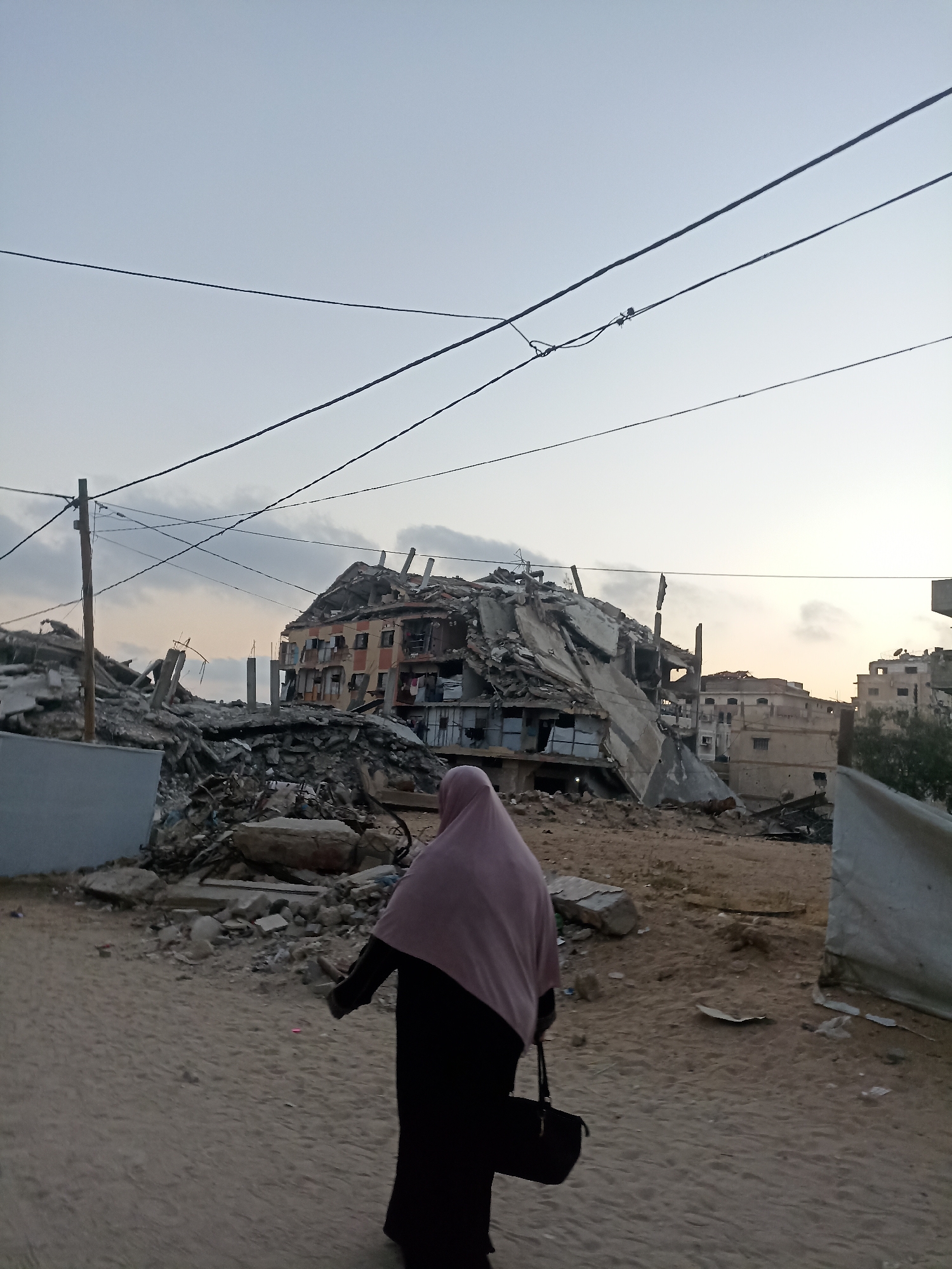 A person wearing a dark abaya and a light pink hijab walks away from the camera across a sandy, debris-strewn area. They are carrying a dark handbag. In the background, a large building has been severely damaged, with most of its structure collapsed or broken. Parts of the building are still standing, showing multiple floors with visible windows and some exposed internal structures. The scene is one of destruction, with rubble and debris covering the ground.
The sky above is overcast and appears to be either early morning or late evening, indicated by the soft, diffused light. Several dark wires crisscross the sky, connected to a wooden utility pole visible on the left side of the frame. The overall setting suggests a post-conflict or disaster zone. The color palette is muted, dominated by grays, browns, and the pale sky. There is no visible text in the image. The atmosphere is somber, conveying a sense of loss and devastation. The location is identified as Gaza, Palestinian Territories.