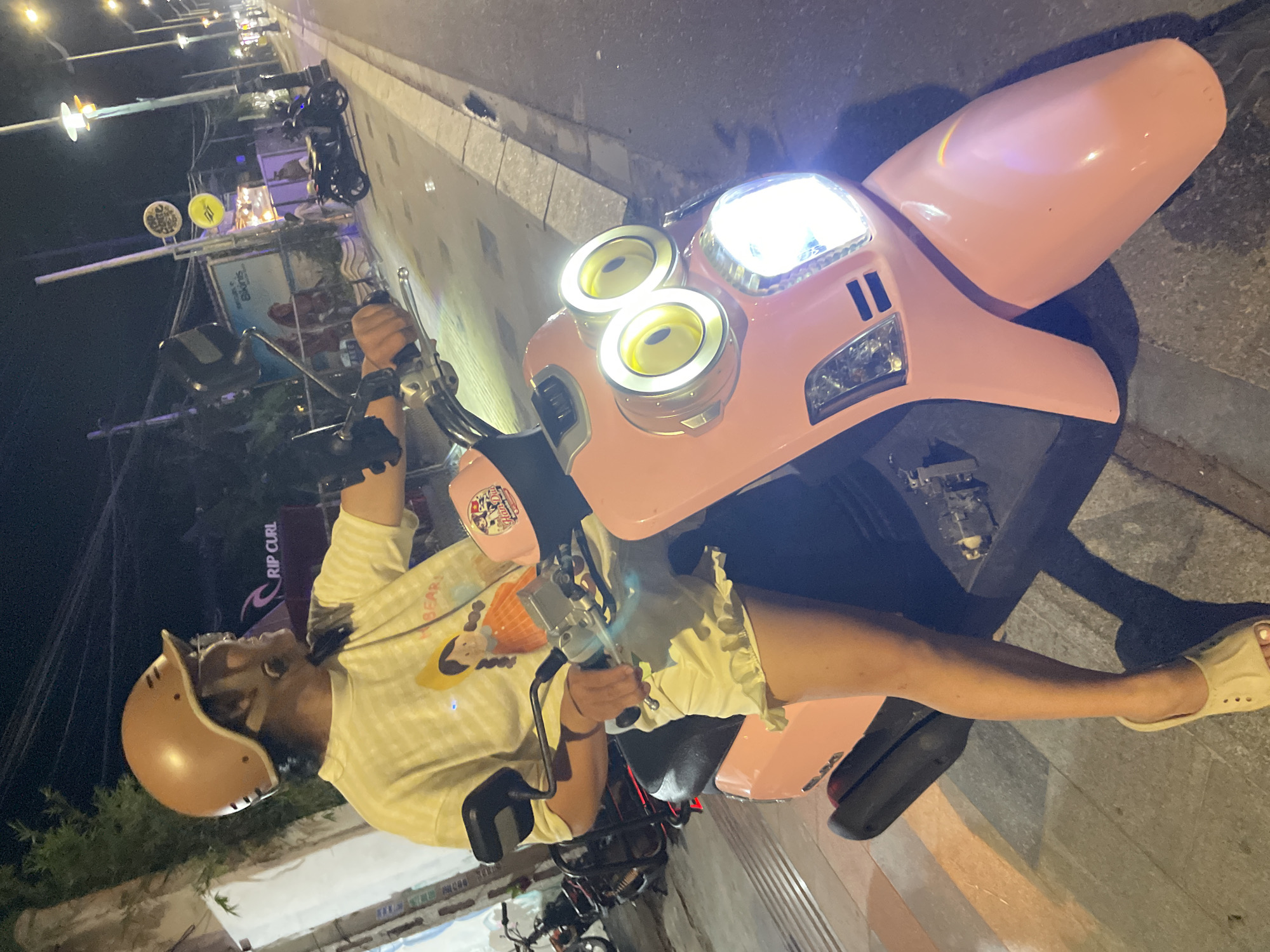 A person sits on a pink scooter at night in Phu Thuy Ward, Vietnam. The scooter has distinctive front lights that resemble the eyes of a Minion character from the "Despicable Me" franchise. The person is wearing a tan helmet, a striped t-shirt with a graphic print, and light-colored shorts. They are holding the scooter's handlebars. The scene appears to be on a street, with streetlights visible in the background, suggesting it is nighttime. The ground is paved, and there are some plants and structures visible along the side of the road. A sign with the text "RIP CURL" is partially visible. The lighting from the scooter's headlights illuminates the front of the vehicle and the person's legs. The overall mood is casual and relaxed.