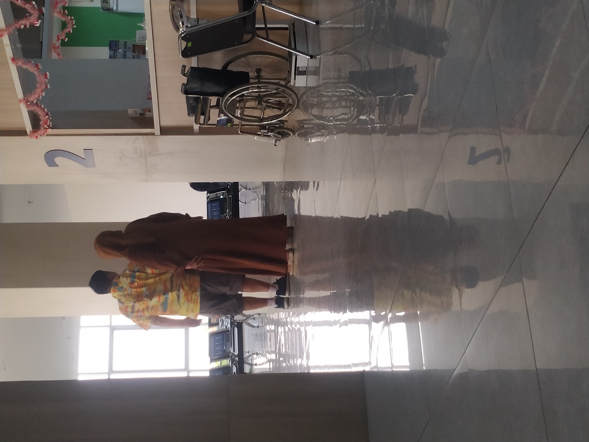 In Mojosongo, Indonesia, two individuals, a man and a woman, stand facing away from the camera in a brightly lit hallway. The man, on the left, wears a colorful, short-sleeved shirt and grey shorts. The woman, to his right, is dressed in a long, brown garment and headscarf. They appear to be conversing or waiting. The hallway features a polished, reflective floor that shows the reflections of the people and the environment. In the background, there are rows of empty chairs and a wheelchair. A large numeral "2" is visible on a pillar. The scene suggests a waiting area, possibly in a medical facility or administrative building.