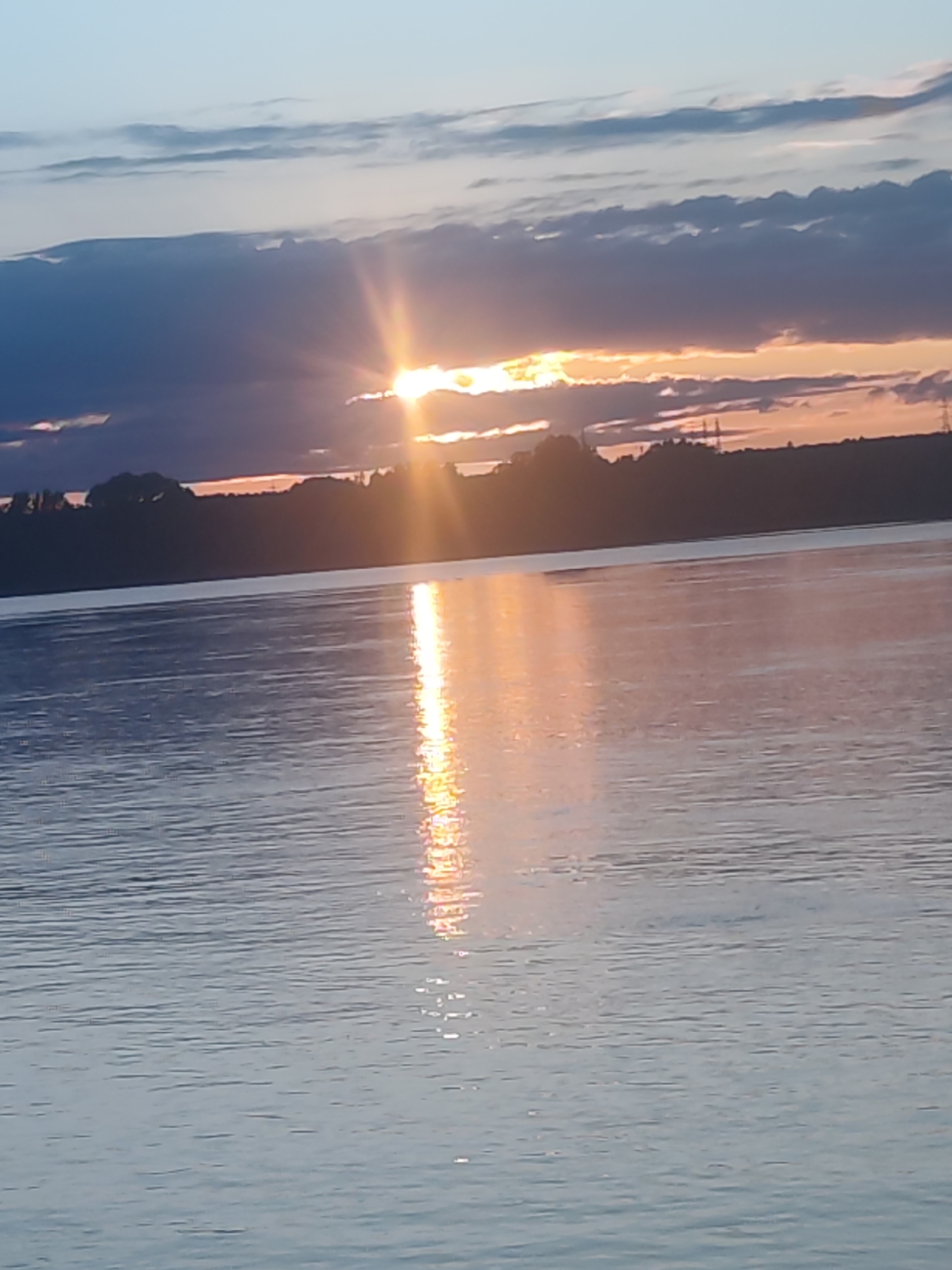 The image captures a serene sunset or sunrise over a wide body of water, likely in Noviy, Russia. The sky displays a gradient from a light blue at the top to warmer hues of orange and yellow near the horizon. The sun, a bright and intense source of light, is partially obscured by dark, heavy clouds, creating a strong sunburst effect.
Its golden light casts a brilliant, shimmering reflection that extends across the calm, rippled surface of the water, creating a striking vertical path of light that contrasts with the cooler blue and grey tones of the surrounding water. Above the darker clouds, lighter, wispy clouds are visible in the upper sky.
On the distant horizon, a dark silhouette of land, likely trees or a distant shore, stretches across. Within this silhouette, faint outlines of man-made structures, possibly electricity pylons, can be discerned, indicating a distant settlement or industrial presence. The scene conveys a sense of peaceful tranquility, characteristic of the golden hour. No people or visible text are present in the image.