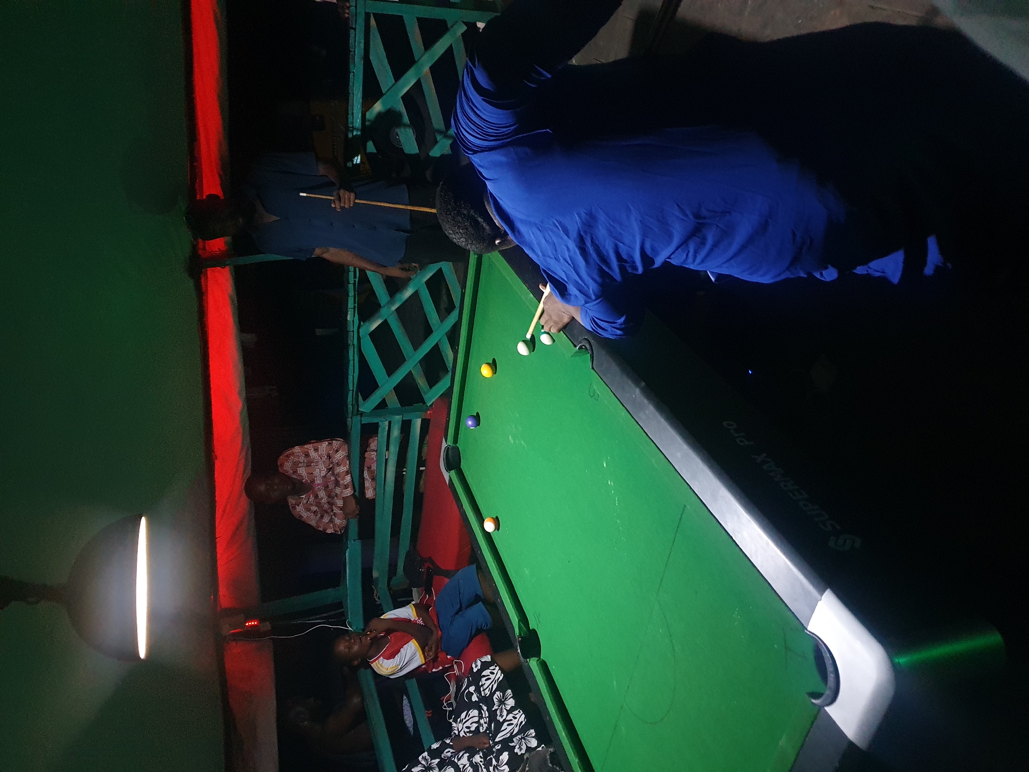 A group of people are gathered around a billiards table in an indoor setting. One person, wearing a blue shirt, is bent over the table, cue in hand, apparently taking a shot. Another person wearing a blue shirt stands on a structure that has a series of X shapes. Several other people are seated and standing near the table, dressed in a variety of clothing. The billiards table is green with balls scattered on the surface. A bright, orb-shaped light fixture illuminates the scene. The environment appears to be a casual gathering space, but specific details of the location in Lokoja, Nigeria, are not discernible from the image.