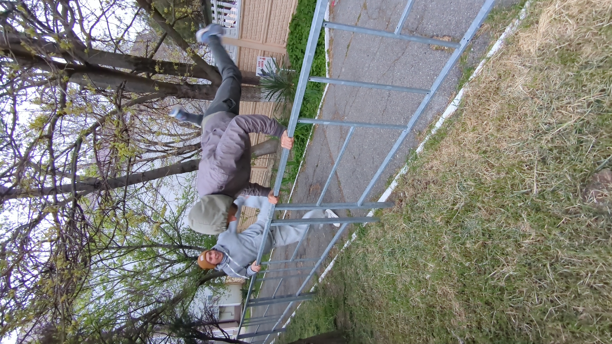 An outdoor, slightly wide-angle shot shows two males engaged in playful or acrobatic activities on a grey metal fence, set against a background of trees, a paved path, grass, and a building. The image is taken on an overcast day, evident from the soft, diffused lighting and light sky.
The taller of the two individuals, wearing a grey jacket and dark trousers with blue sneakers, is inverted, performing a handstand-like maneuver on the top rail of the fence. His hands grip the rail firmly, and his legs are extended upwards, with one foot seemingly resting against a tree trunk or branch.
Below him, a younger male, dressed in a grey hoodie, light grey pants, and white sneakers, with a brown beanie, hangs from a lower horizontal bar of the same fence. He faces the camera, his feet off the ground, and displays a clear smile, indicating enjoyment.
The setting appears to be a public or residential area. To the left, a mature tree with budding light green leaves, characteristic of spring, dominates the background. To the right of the fence, a strip of dry and green grass is visible, separating it from a paved, asphalt-like path or road. Further back, a light-colored building with horizontal siding can be seen, partially obscured by green foliage and a small palm-like plant.
A small, rotated sign is visible on the building or near the plant in the mid-ground. The text "АЛЫЕ ПАРУСА" (Alye Parusa), which translates to "Scarlet Sails," can be discerned on the sign. This is a common name for hotels or establishments in Russia, particularly in coastal areas like Gelendzhik. The overall mood of the image is lighthearted and energetic.