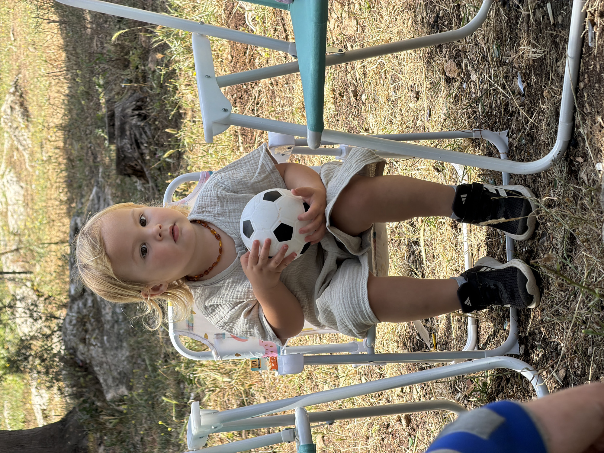 In an outdoor setting, a fair-skinned toddler with blonde hair sits in a highchair, holding a small, black-and-white soccer ball. The child is wearing a light-colored, loose-fitting outfit and dark sneakers with white stripes. A brown beaded necklace adorns their neck. The highchair is white with a light blue and green support bar. The ground is covered in dry grass and leaves. In the bottom right corner, part of a person's leg wearing blue shorts is visible. The location is suggested to be in the Zvulun Regional Council, Israel.