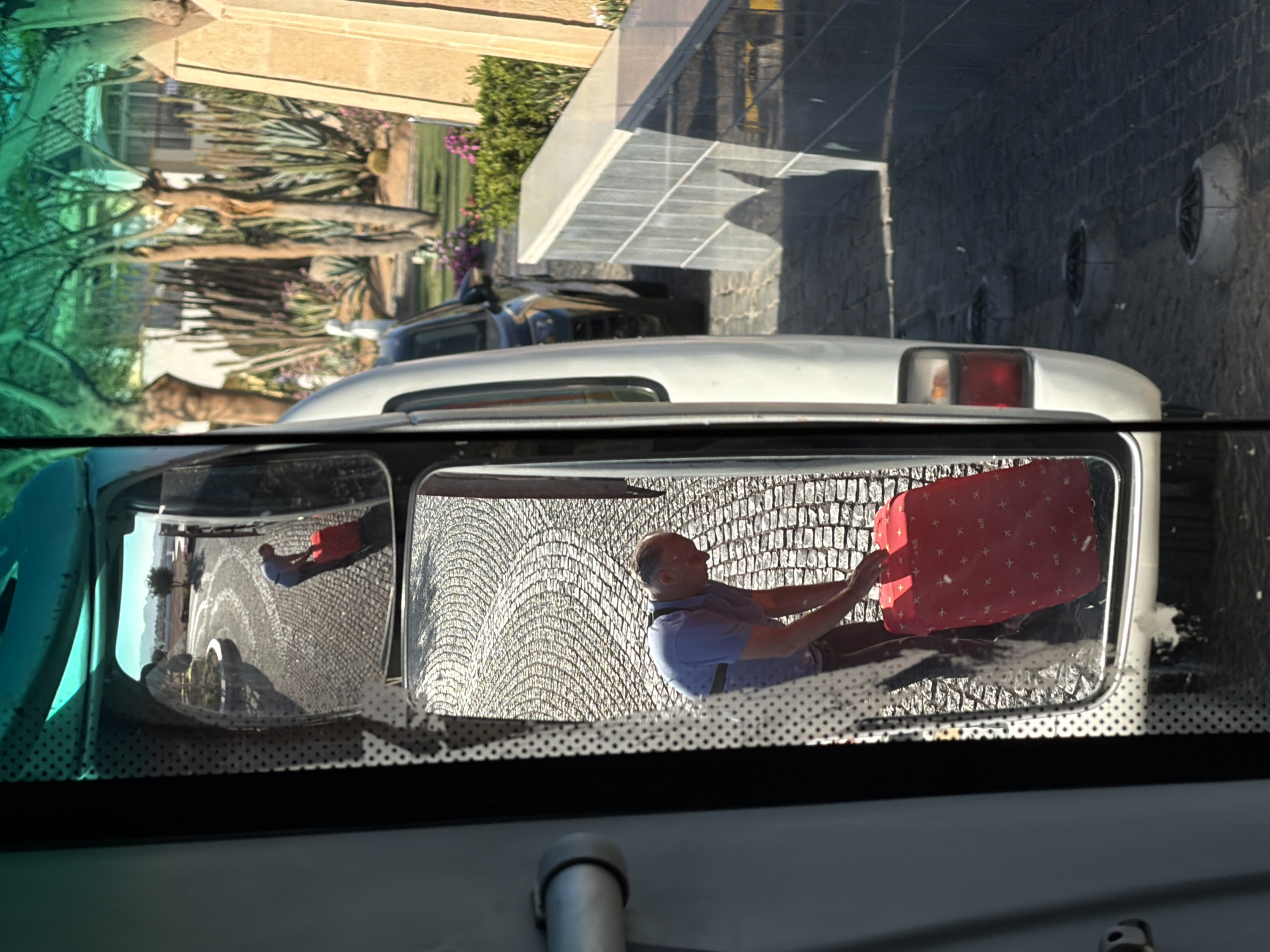The image captures a scene from the interior of a vehicle, looking outwards through the windshield and focusing on the side-view mirror. The overall setting is brightly lit by sunlight, indicating daytime with clear weather.
In the prominent side-view mirror, a man is reflected. He has short dark hair and is wearing a blue polo shirt with dark straps (possibly suspenders or part of a uniform) and dark trousers or shorts. He is actively engaged with a red suitcase, which features a pattern of small white cross-like or airplane symbols. He appears to be either loading, unloading, or adjusting the position of the suitcase, holding it with both hands. The ground reflected in the mirror is paved with distinctive light-colored, textured cobbles or bricks, arranged in a wavy pattern, suggesting a stylized driveway or path.
Outside the mirror's reflection, to the left (through the green-tinted windshield), mature tree trunks and green foliage, consistent with palm trees or desert flora, are visible. Part of a light-colored building is also discernible. To the right, beyond the vehicle's white roofline, is a modern building entrance with a transparent or light-colored angular canopy, casting distinct shadows on the dark grey, textured wall below. Several round, possibly decorative or ventilation, fixtures are mounted on this wall. The white rear section of the vehicle (likely a car or van) is visible, including a red taillight.
The scene suggests an arrival or departure at a building, possibly a hotel or resort, given the man handling luggage and the architectural style. There is no visible text in the image. The location context provided indicates Sharm El Sheikh, Egypt, which aligns with the observed architecture, vegetation, and sunny conditions.