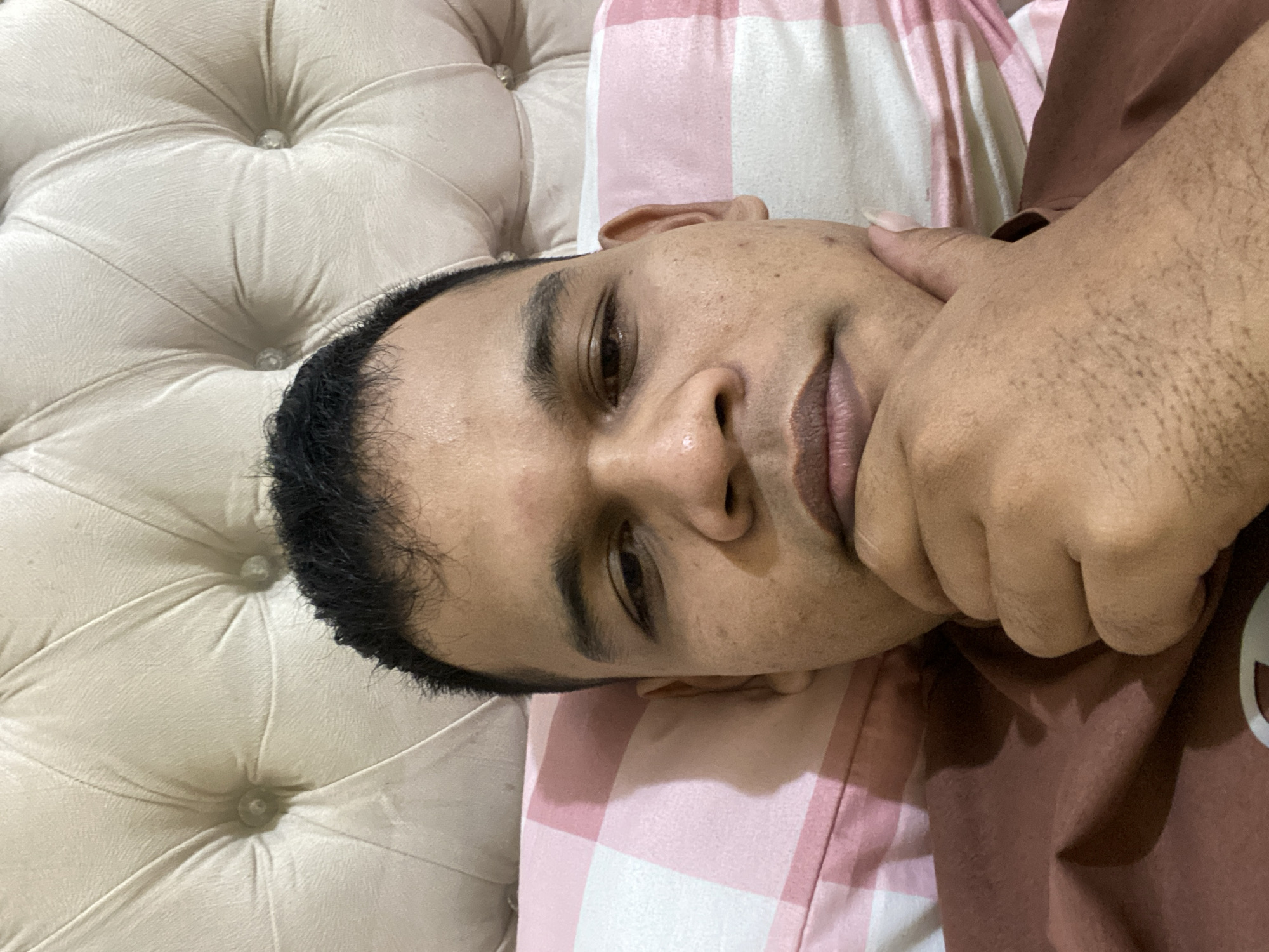 A close-up, self-taken portrait shows a person lying in bed, with their head resting on a pink and white checkered pillow. The background features a beige, tufted headboard. The person is looking directly at the camera with their chin resting on their left hand. They are wearing a brown shirt. The scene appears to be indoors, likely in a bedroom in Lambaro Kueh, Indonesia.