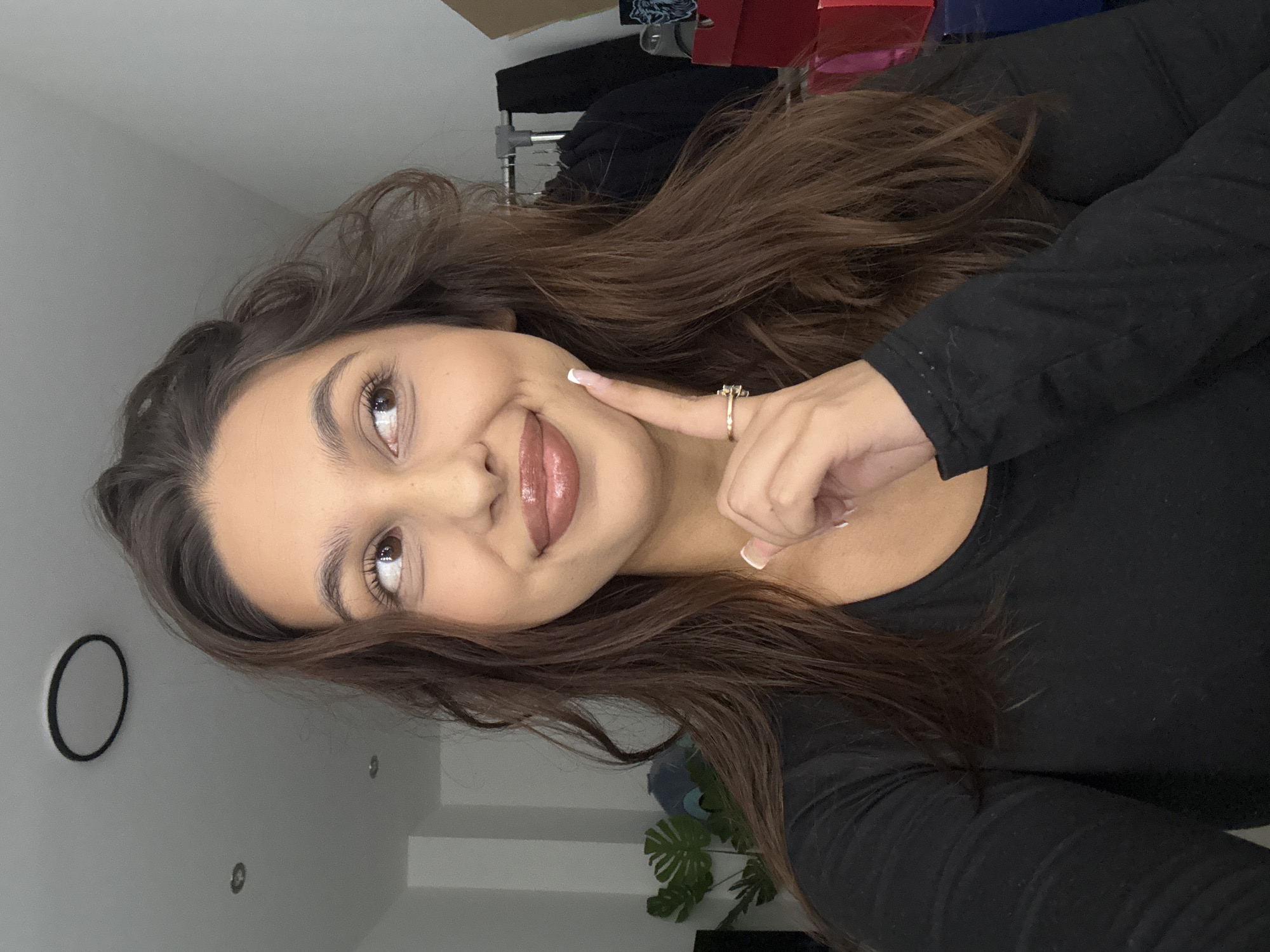 A close-up portrait of a young woman, taken indoors in what is contextually identified as Mönchengladbach, Germany. The image is rotated 90 degrees clockwise, showing the woman's head and upper torso. She has long, dark brown, wavy hair, brown eyes, and full lips adorned with glossy nude or pink lipstick. She wears a black long-sleeved top. On her right hand, she sports a gold-colored ring with small stones, and her fingernails feature a French tip design.
She is looking upwards and slightly to the right with a soft, playful smile. Her right index finger is gently touching her right cheek, creating a slight dimple. The background is primarily a plain light gray wall, suggesting a simple indoor setting, possibly a room in a home. A black circular object is visible on the wall to the left. In the upper right background, some red and dark-colored boxes or objects are partially seen, hinting at storage or shelving. A hint of green foliage from an indoor plant is visible in the lower right corner. The lighting is bright and even, indicating it is likely daytime. There is no visible text in the image. The overall impression is one of contentment and playfulness.