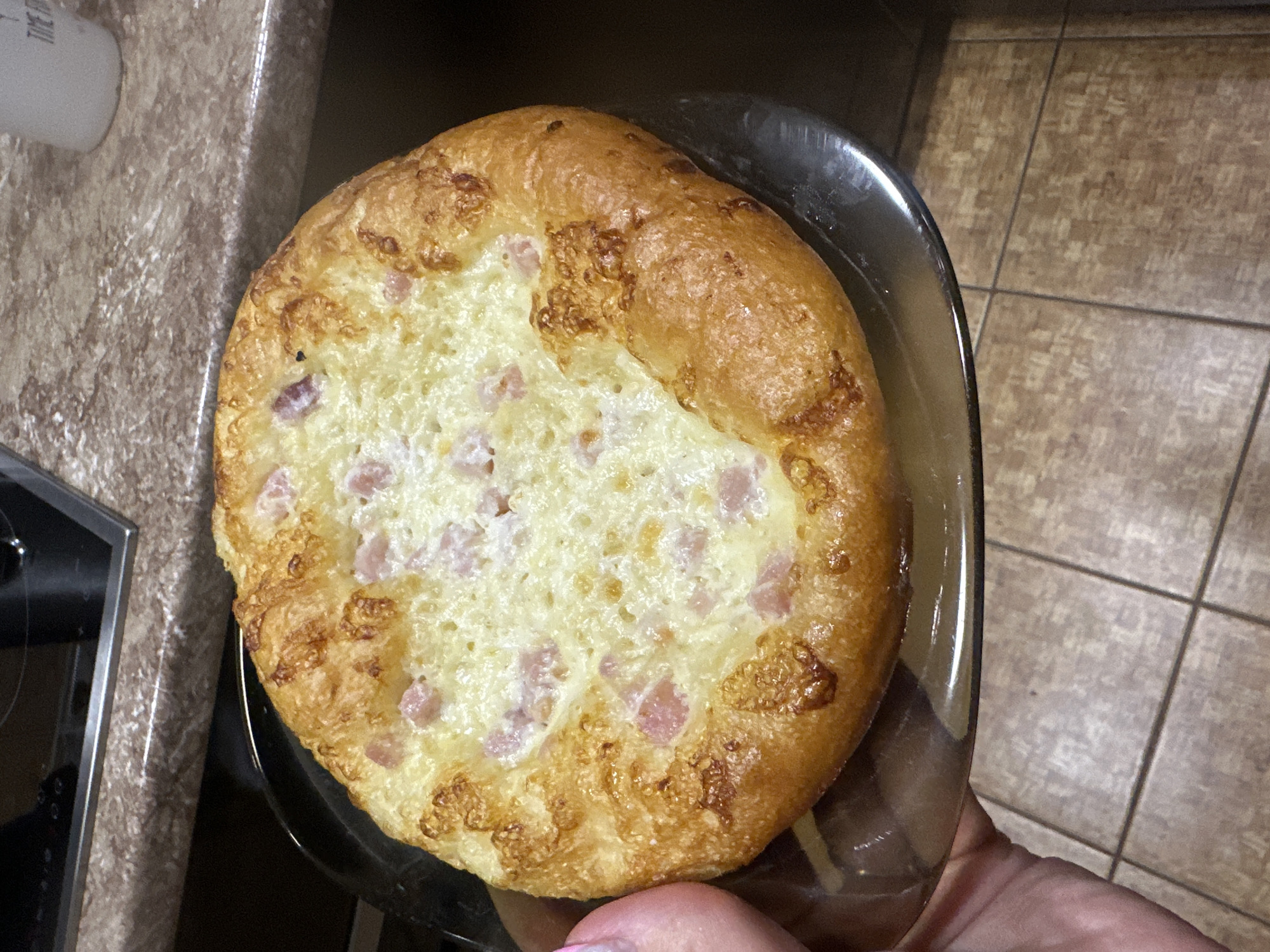A round, golden-brown baked good, likely a pastry or small pizza, sits on a dark glass plate. The pastry has a raised crust and a creamy, melted cheese topping with small pieces of pink ham visible. A hand with a pink fingernail is holding the plate from the left side of the image. The background appears to be a kitchen countertop with a speckled brown and gray pattern, and a glimpse of a black stovetop is visible at the top left. The floor below is tiled with a light brown, textured pattern. The location is identified as Sayanogorsk, Russia.