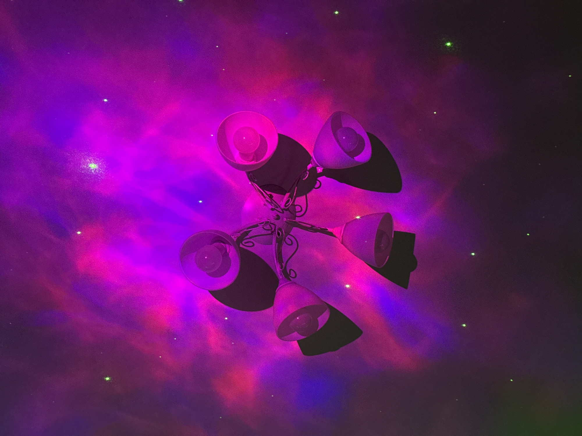 The image presents a low-angle perspective of a multi-bulb lamp fixture. The lamp has five individual shades, each containing a light bulb. The shades are oriented in different directions. The scene is illuminated with a pink and purple light, giving the appearance of a nebula or galaxy. The backdrop is a dark space filled with scattered green spots, resembling stars. The location in the city of Unknown, Unknown is indeterminable.