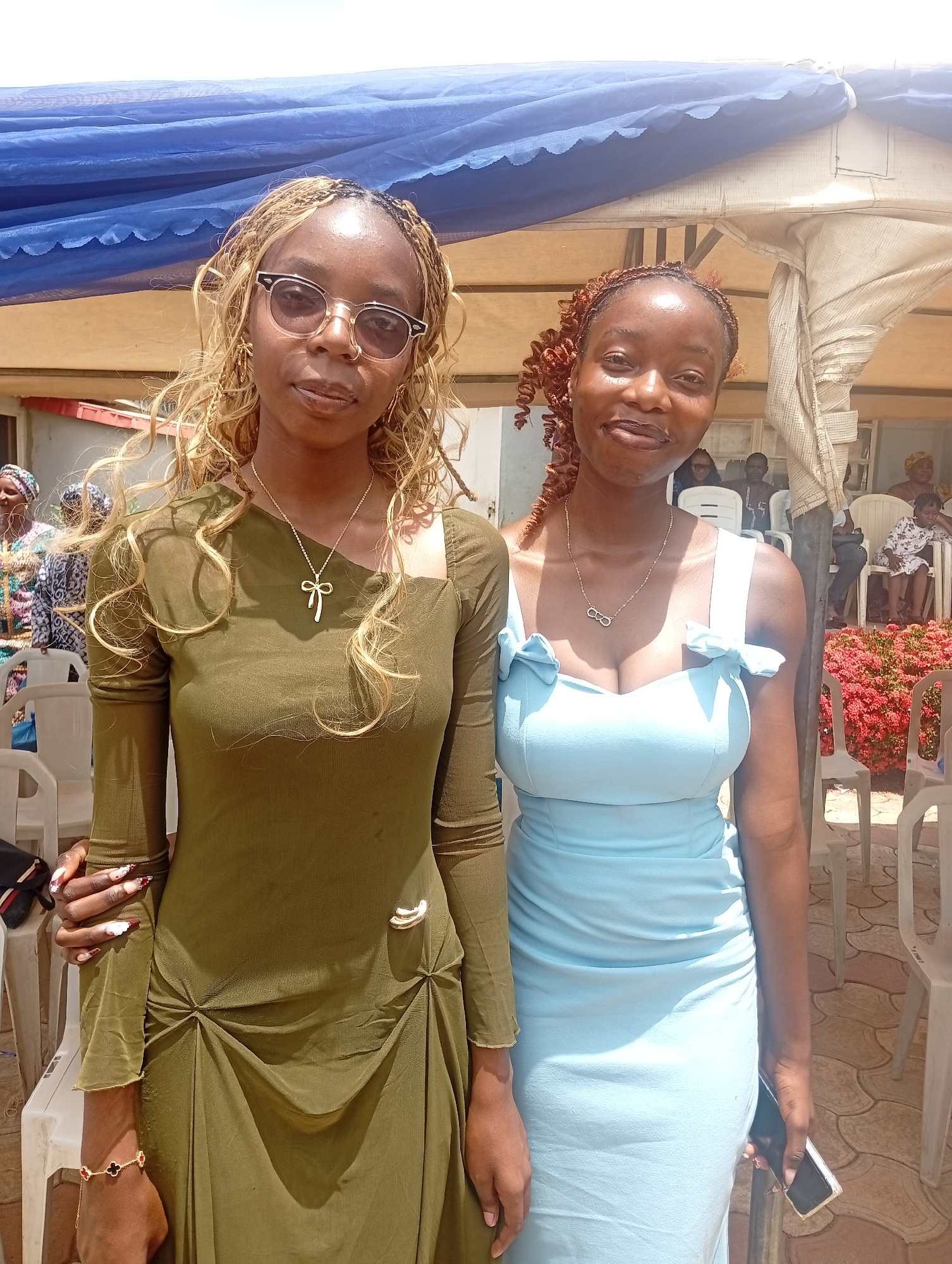Two young women are posing for a photo outdoors, likely at an event. The woman on the left has blonde braided hair and is wearing glasses and a green dress. She has a gold necklace with a bow pendant and a bracelet with a clover charm. The woman on the right has curly reddish-brown hair and is wearing a light blue dress with bow details on the shoulders. She has a silver necklace with an infinity symbol pendant.
They are standing in front of a blue tarp and a tent-like structure, with a background of people sitting in white plastic chairs and some red flowers. The lighting suggests it is daytime, and the weather appears sunny. The ground is paved with interlocking tiles. The presence of chairs and people in the background indicates a gathering or event. The overall atmosphere seems relaxed and festive. The context of Barden Yana, Nigeria, suggests a possible cultural event or celebration. There is no visible text in the image.