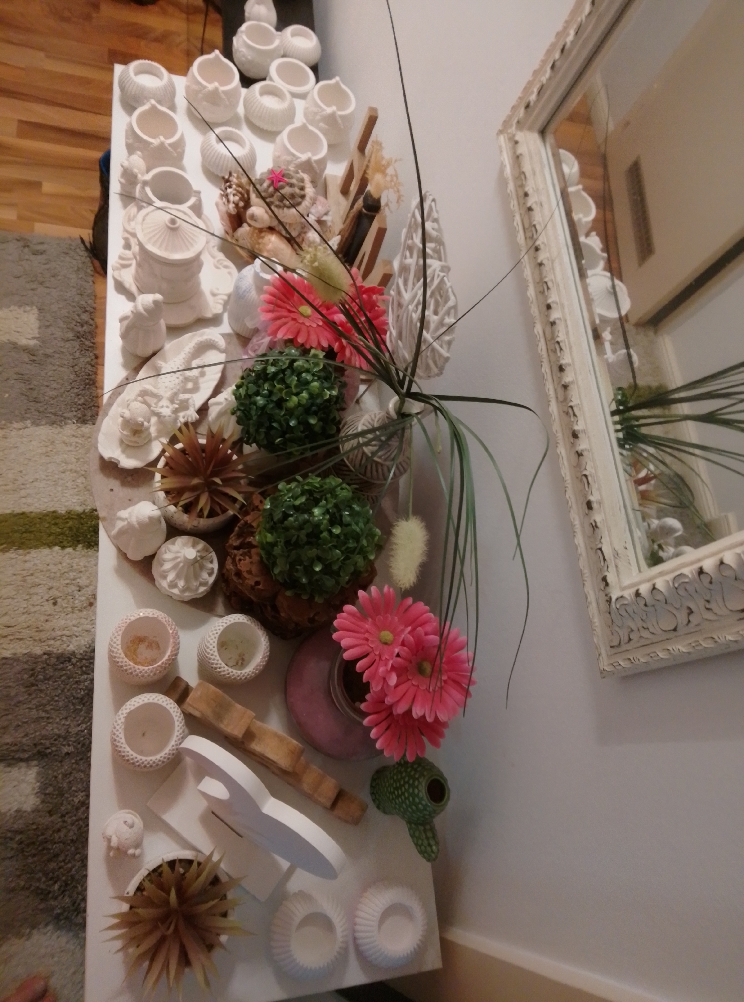 The image shows a white table with various decorative items. On the left side of the table, there are several white ceramic pots of different sizes and shapes, some with decorative patterns. There are also some white ceramic figurines, including a small bird and a lizard. In the center of the table, there is a collection of items including a pink artificial flower arrangement, two green artificial topiary plants, and a brown decorative object that appears to be a dried plant or wood. To the right of the table, there is a large, ornate white mirror. The table is placed on a floor with a rug that has stripes of gray, brown, and green. The background shows a wooden floor and a wall. The overall scene appears to be an indoor decorative arrangement, possibly in a home or a shop. The location provided, Wedel, Germany, does not appear to be directly depicted in the image itself, nor is there any context within the visual information to confirm this specific geographical placement. The image captures a static display of various decorative objects.