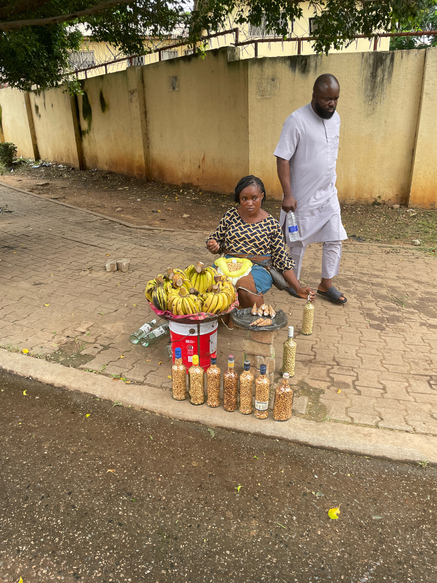 A man in a light grey traditional outfit and a woman in a patterned top are on a paved street next to a yellow wall with barbed wire on top. The woman is sitting on the ground next to a large red and white container filled with bananas. In front of the container are several bottles filled with nuts, and to the left of the bananas are small fried fish. The man is standing and holding a bottle of water. The ground is wet and has scattered leaves and debris. The scene appears to be a street market in Abuja, Nigeria.