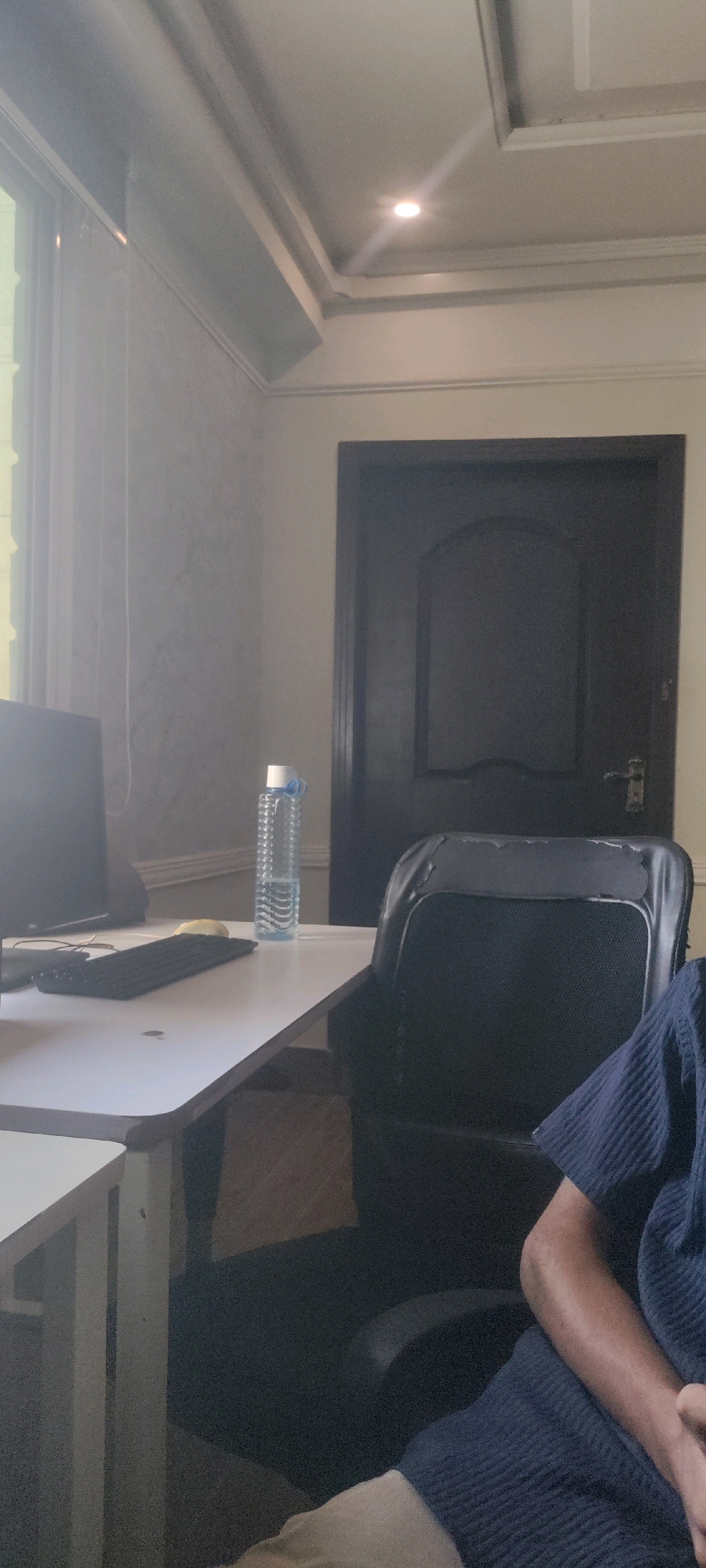 The image depicts an indoor scene, likely an office or study, in Khana Dak, Pakistan. A desk with a computer monitor and keyboard is visible on the left, with a water bottle placed on the desk. A black office chair is positioned behind the desk, showing signs of wear. In the background, a dark wooden door with a handle is present. The ceiling features recessed lighting and decorative molding. The overall lighting suggests daylight filtering in from a window, casting a slightly hazy effect. A portion of a person, wearing a blue shirt and beige pants, is visible in the foreground on the right, seated in or near the chair.