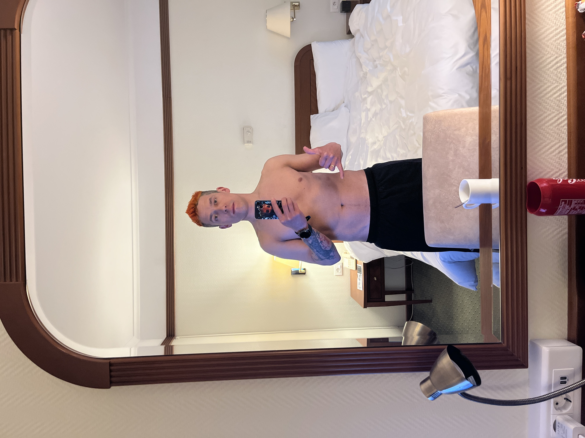 A bare-chested man with reddish-orange hair is reflected in a large, ornate mirror. He holds a phone in his left hand, seemingly taking a selfie. A tattoo is visible on his left arm. He is wearing black shorts. The reflection shows a hotel room with a bed, a nightstand, a white mug, and a can of Coca-Cola on a surface in front of the mirror. The wall behind the bed is white with a patterned texture. The overall setting appears to be a hotel room, possibly in Moscow, Russia. A bedside lamp with a flexible neck and a modern design is mounted on the wall near an electrical outlet.