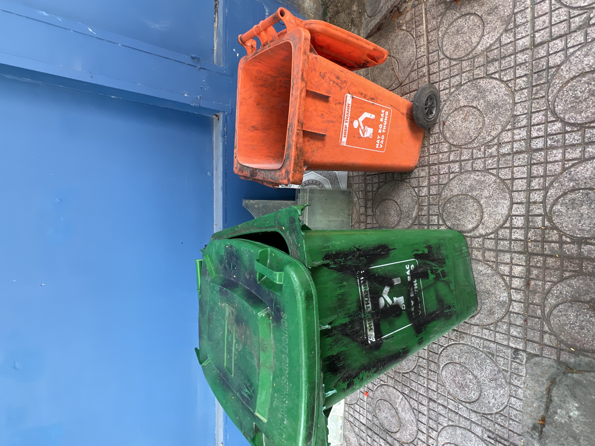 An exterior medium shot captures two plastic trash bins placed on a tiled sidewalk in front of a blue wall. The trash bins are a vibrant green and orange. The green bin is larger, with a hinged lid that is partially open. Both bins appear weathered and have black graffiti markings on their sides. The orange bin has a single wheel attached to its base and a flip-up lid. The tiled sidewalk has a repeating circular pattern. The overall scene suggests an urban environment in Nha Trang, Vietnam, likely a street or public area.