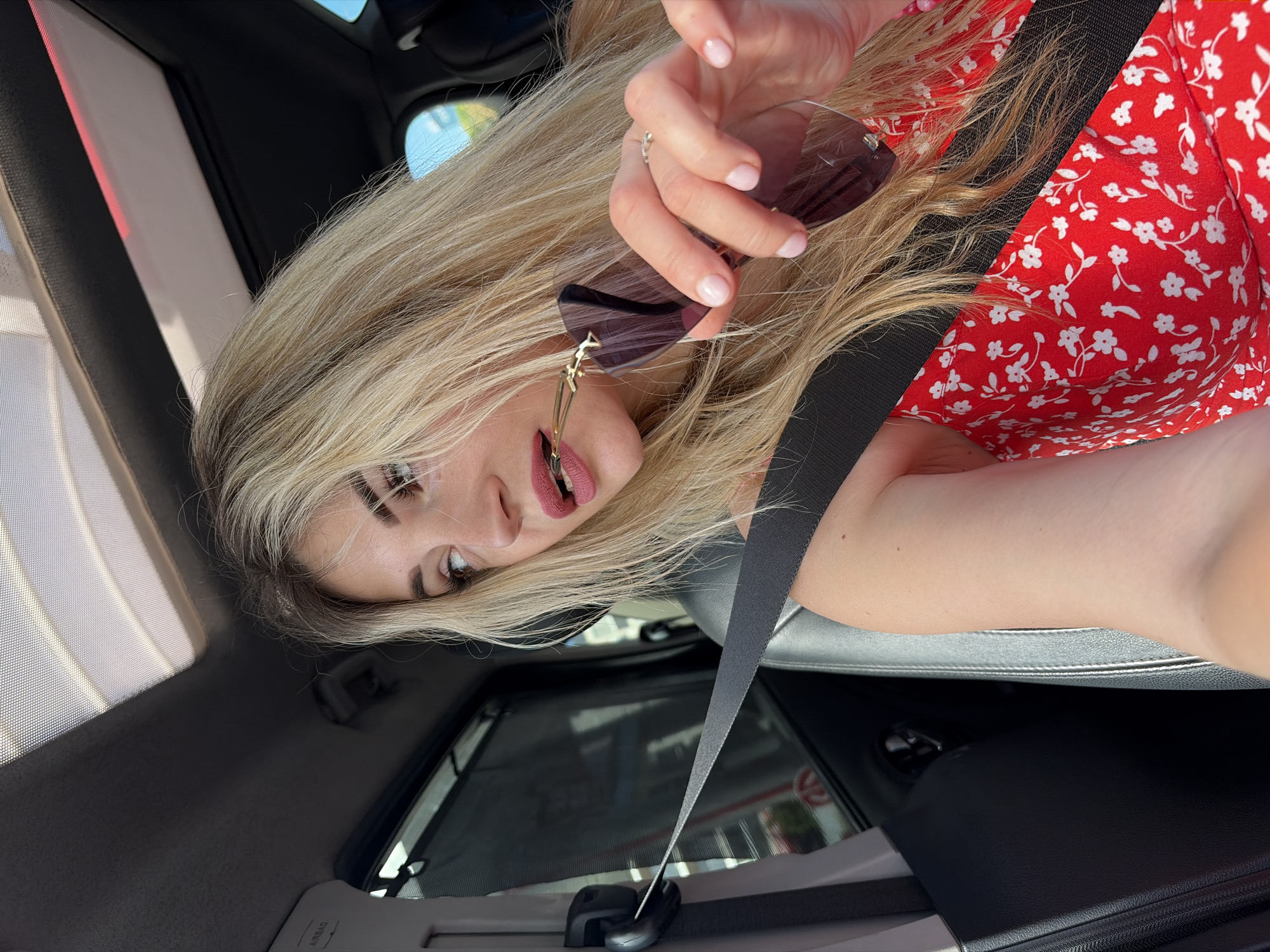 A blonde woman is depicted inside a vehicle, occupying the front passenger seat in the city of Podgorica, Montenegro. She is secured by a black seatbelt stretching diagonally across her chest. Her long, light-colored hair frames her face and extends over her shoulders and chest. She is wearing a red dress with a white floral pattern. Her lips are colored with pink lipstick, and she is biting the golden arm of a pair of dark-lensed sunglasses, which she holds in her right hand. Her fingernails are painted with a light pink polish, and a ring is visible on her right ring finger. Her gaze is directed towards the right side of the frame, looking out the car window. The vehicle's interior features dark upholstery, including the headliner, seats, and door panels. An open sunroof is visible overhead, partially covered by a fine mesh screen. An "AIRBAG" label is affixed to the upper left pillar. A dark mesh sunshade is deployed over the rear passenger window. A subtle red ambient light strip is present along the right edge of the sunroof trim.