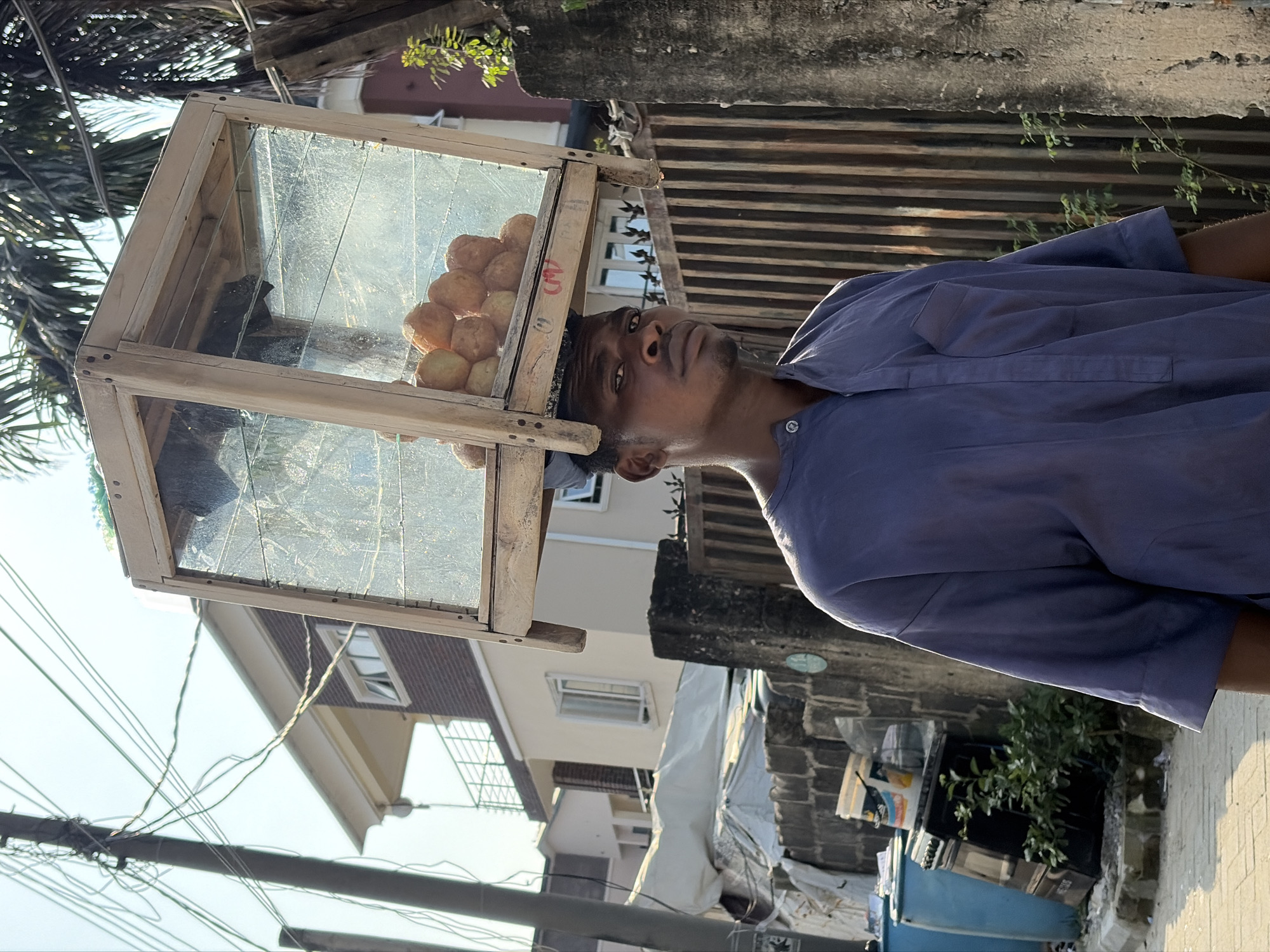 The image shows a young man in a blue shirt with a wooden and glass display case balanced on his head. The display case contains several round, golden-brown food items, likely pastries or fried dough. The man is looking upwards and slightly to his left, with a neutral expression. The scene is outdoors, with a building in the background and power lines visible in the upper left quadrant. The overall lighting suggests it is daytime, possibly late afternoon given the shadows. The setting appears to be a street or market area in Igboefon, Nigeria, as indicated by the style of the buildings and the presence of street vendors. There is no visible text in the image.
