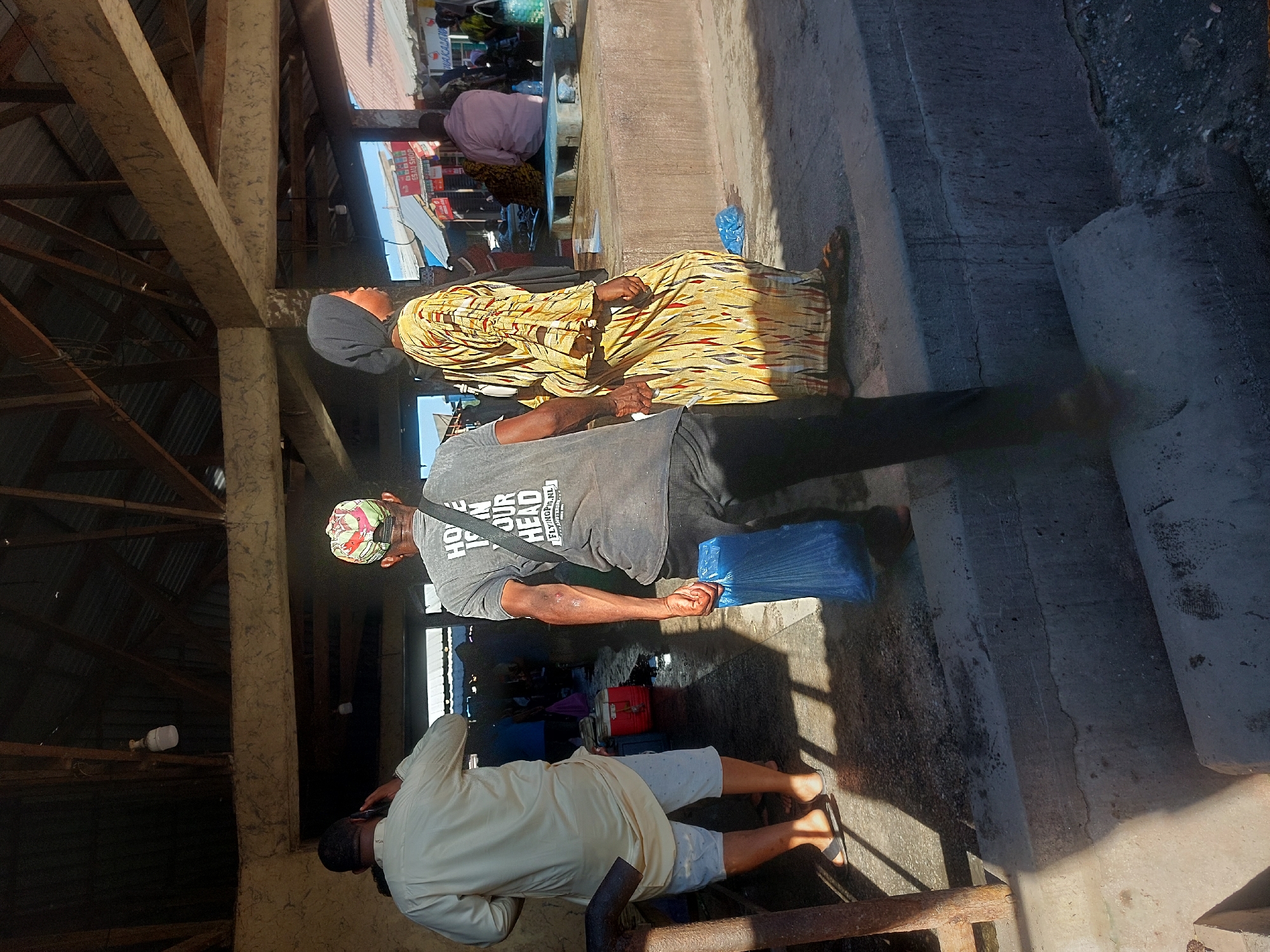 The image shows an outdoor scene in Dar es-Salaam, Tanzania. A man in a grey shirt and black pants holds a blue plastic bag. He's walking on a concrete surface near steps. A woman in a yellow patterned dress walks beside him. Another person in a light-colored shirt and shorts is also present, standing near a metal structure. In the background, there is a view of a busy street market under a roof. The image is taken with the camera in landscape and has been rotated 90 degrees.