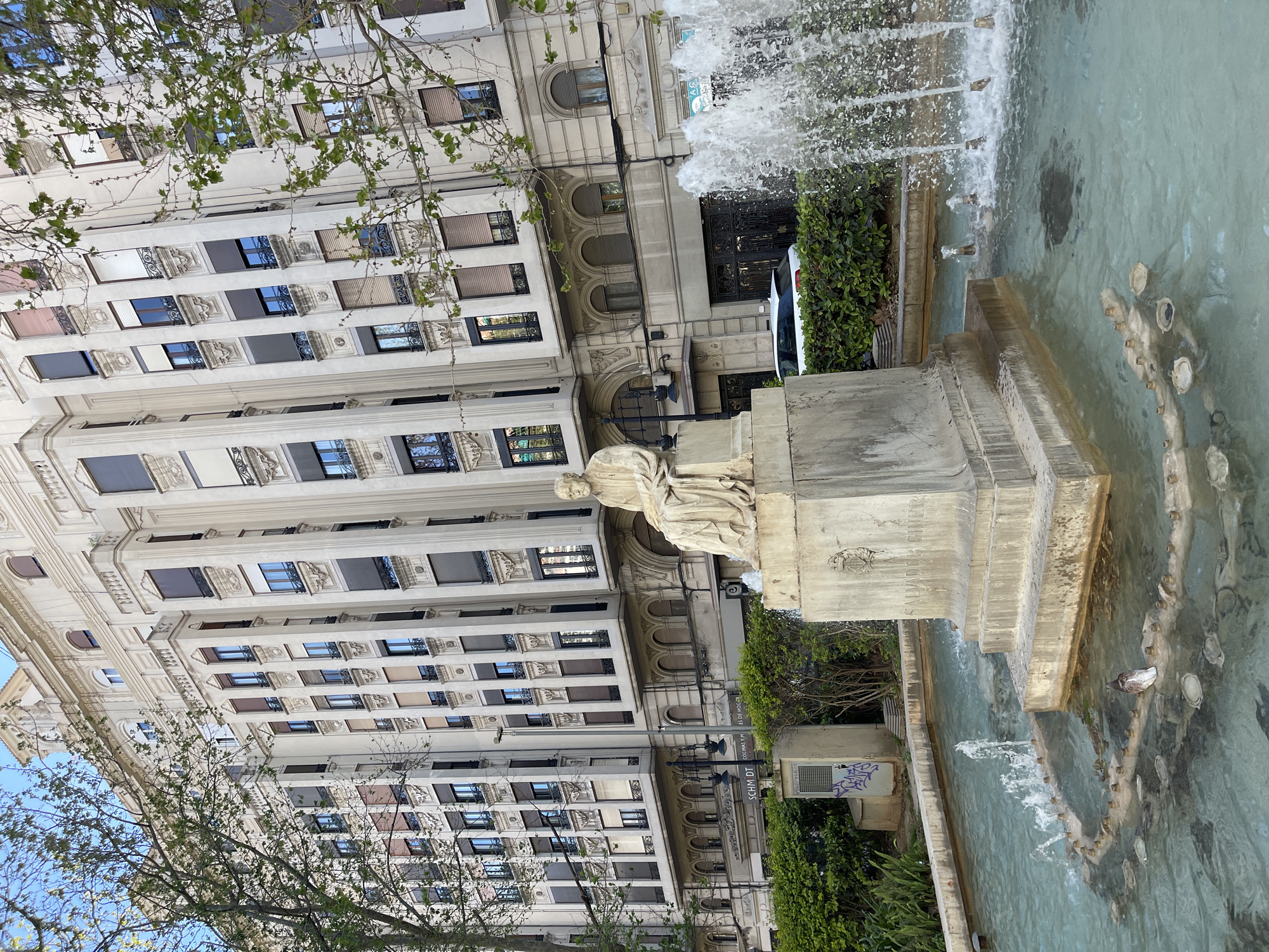 A vertical shot shows a statue atop a plinth beside a fountain, with a light-colored building in the background under a blue sky with tree branches in the foreground. The fountain is shooting water, and the water appears teal. A pigeon is visible in the fountain. The statue appears to be of a robed woman. The light-colored building has many stories of windows, some covered by dark window shades. The plinth has an inscription on it in Spanish. The building is the Finca de Hierro, located in the city of València, Spain.
