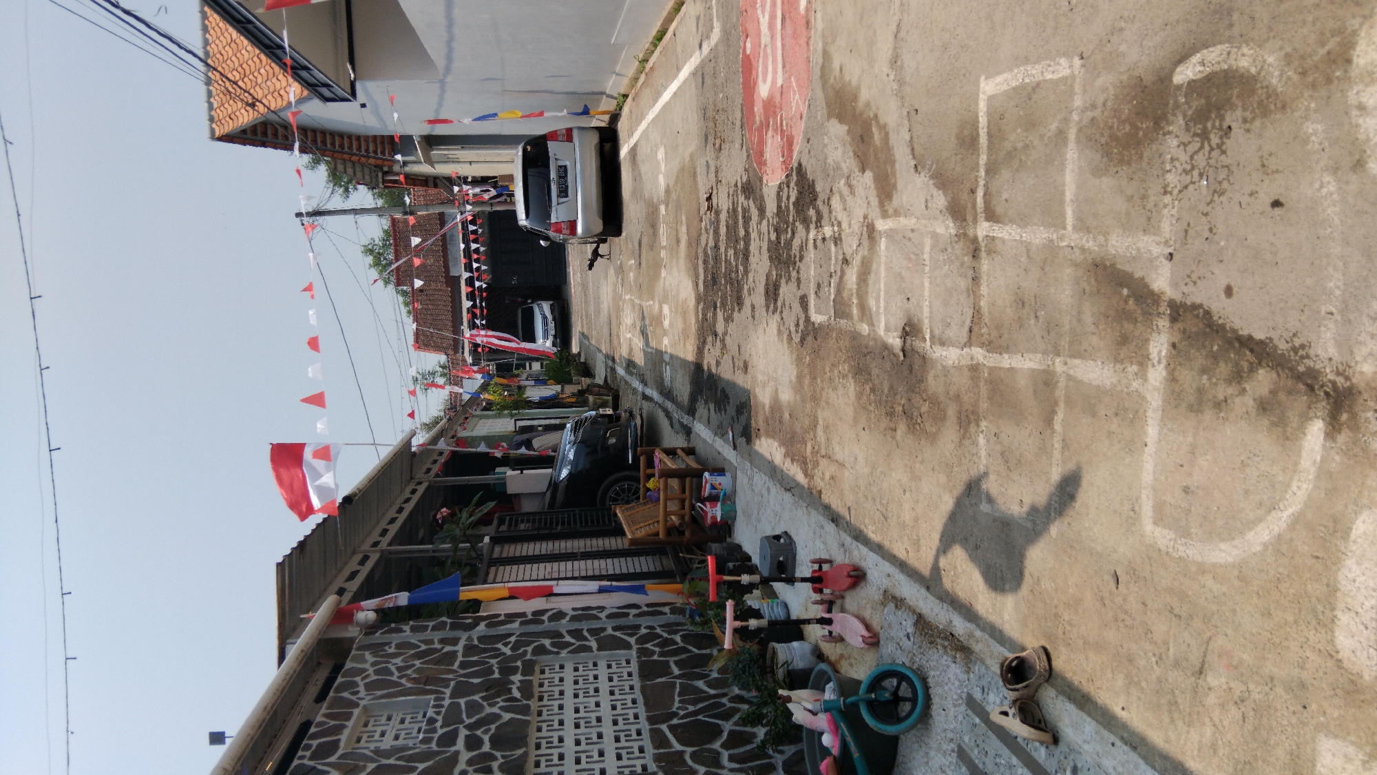 The image depicts a narrow, paved alleyway in the city of Pasekaran, Indonesia. The alley is flanked by buildings, one on the left with a stone facade and another on the right with a white wall and orange-tiled roof. Decorations of red and white triangular flags are strung across the alleyway, suggesting a celebration or national holiday.
On the left side of the alley, a black car is parked, partially obscured. Several children's toys are visible on the ground, including a blue and black scooter, a pink scooter, and a blue tricycle. A wooden bench sits near the stone-walled building.
In the center of the alley, a silver car is parked, and a dark-colored animal, possibly a cat, is walking on the pavement. White lines are painted on the ground, forming a hopscotch grid and a circular target.
The ground is a mix of concrete and dirt, with some wet patches and debris. The sky is clear and bright, indicating daytime. The overall impression is of a residential area decorated for a festive occasion.
