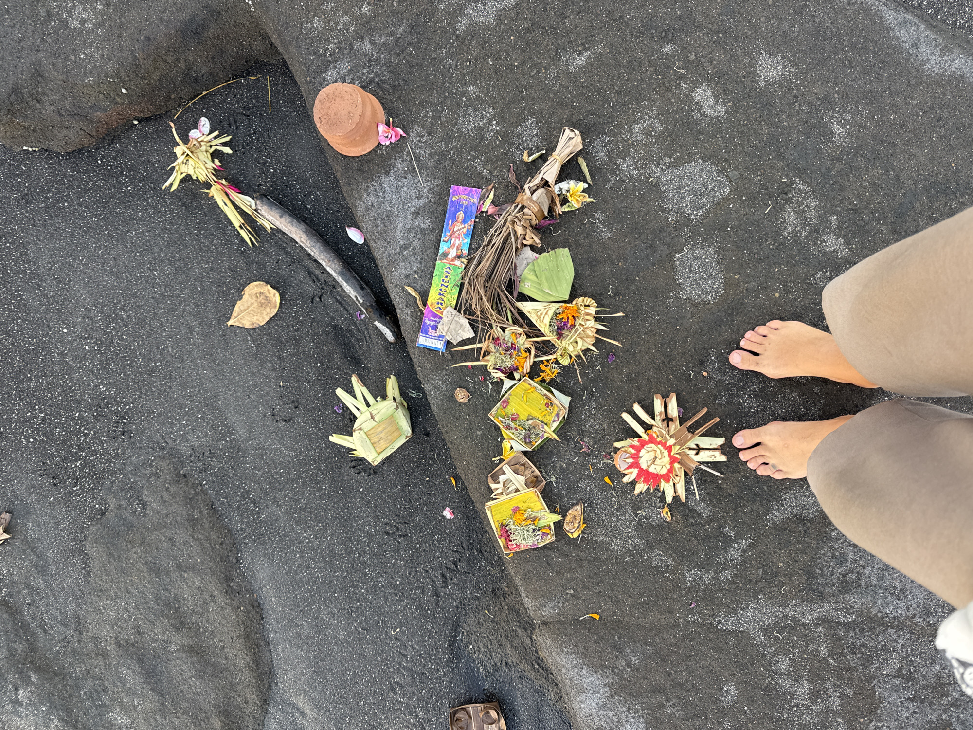The image shows a top-down view of offerings placed on dark, rocky ground, likely a beach or coastal area. The offerings appear to be Balinese canang sari, consisting of woven palm leaf baskets filled with flowers, rice, and other decorative elements. There are several of these offerings scattered across the ground, some with incense sticks and a small terracotta pot. The ground is a dark, speckled texture, possibly volcanic sand or dark rock. In the lower portion of the image, a person's bare feet and lower legs are visible, wearing light-colored shorts. There is no discernible city of Kediri context in the image.