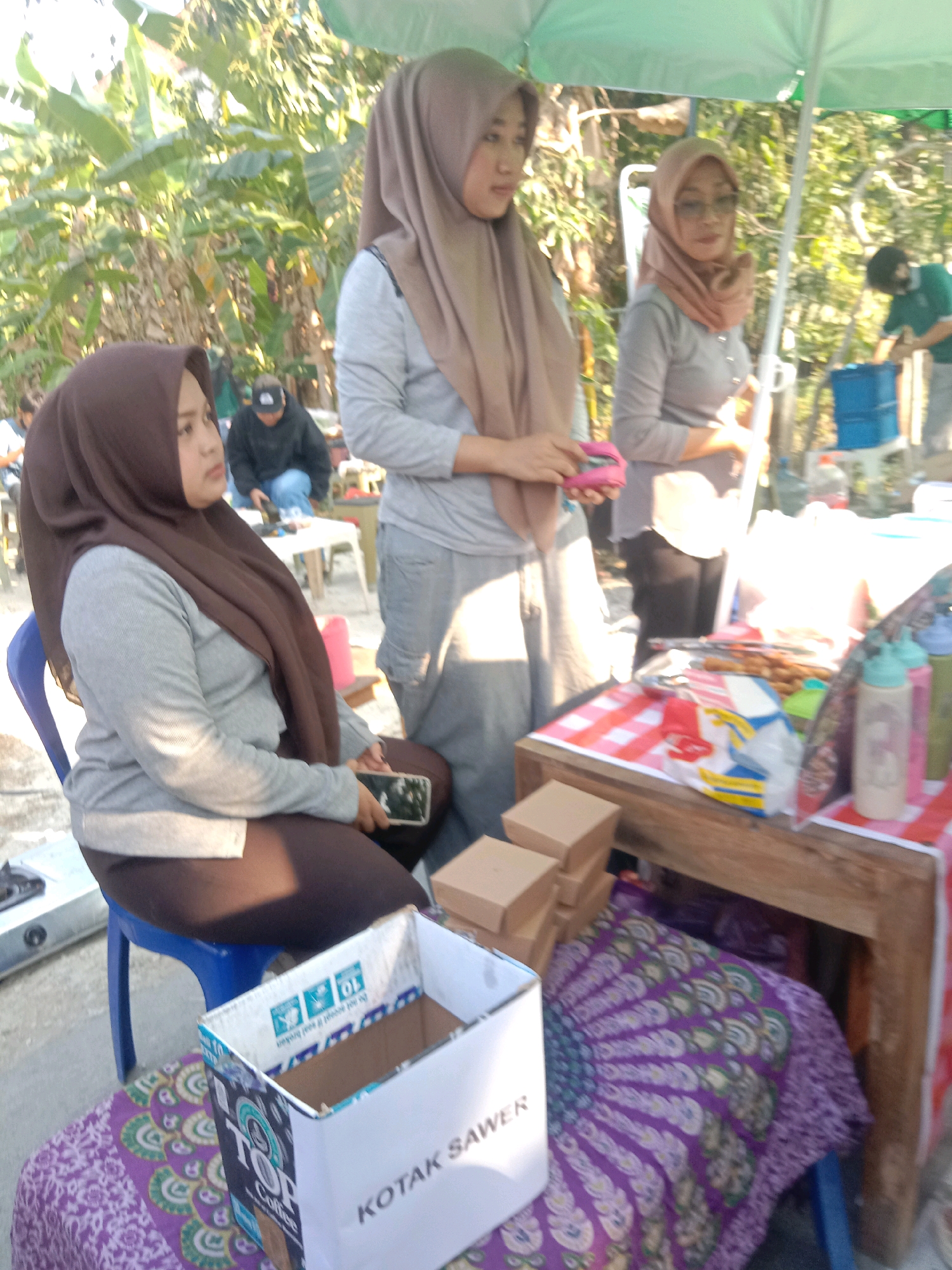 In Banjardowo, Indonesia, a group of individuals are gathered outdoors near a stall, possibly for a market or a food vendor. The scene is bathed in natural sunlight, with lush green foliage framing the background. Three women, all wearing hijabs, are the central figures. One woman, seated on a blue chair, is looking down at her phone, which is resting on her lap. She is wearing a brown hijab and a light gray long-sleeved shirt, with dark brown pants. To her right, another woman stands, also wearing a brown hijab and a light gray long-sleeved shirt, holding a pink object. Further to the right, a third woman, in a lighter brown hijab and a gray long-sleeved shirt, is looking towards the right side of the frame. In the background, a person in a black hoodie and a person in a green shirt are visible, engaged in activities near a table. The stall features a wooden counter covered with a red and white checkered cloth, displaying various items including snacks, bottles, and packaged goods. A white box with text reading "KOTAK SAWER" is also prominent in the foreground, alongside stacked cardboard boxes and a patterned purple cloth spread on the ground.