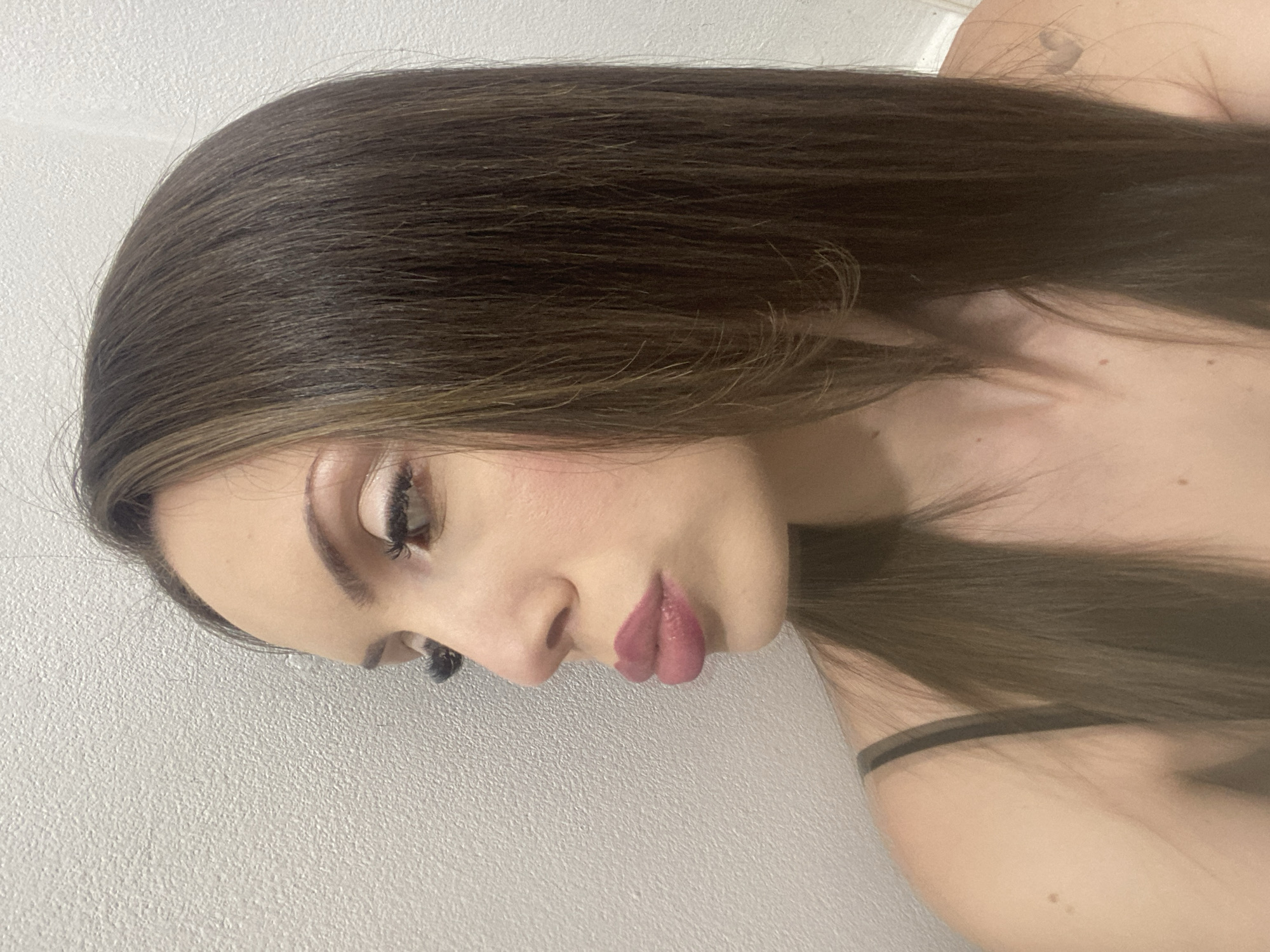 A medium shot shows a woman in profile facing left, with her head tilted slightly down. She has long, dark brown hair with subtle highlights, and her head is close to a textured white wall. She is wearing a dark-colored tank top, and a faint tattoo of a heart is visible on her right shoulder. Her face is made up, with noticeable eyeliner and mascara on her lashes, and her lips are painted a matte pink color. She appears to be pursing her lips. The lighting is soft, and the background is plain, suggesting an indoor setting. The specific location within "Unknown, Unknown" is not discernible from the image.