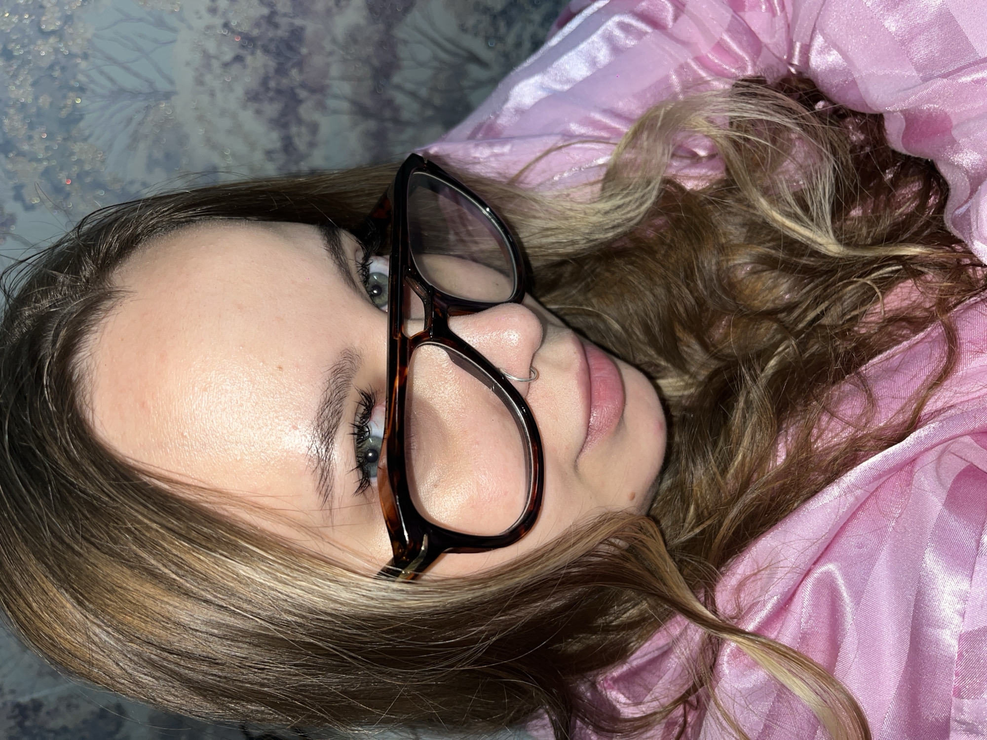 A person with light brown hair, wearing tortoiseshell glasses and a small nose ring, is lying down against a backdrop of pink fabric. The location is not discernible from the image and there is no information to suggest this photo was taken in Novopokrovskaya, Russia.