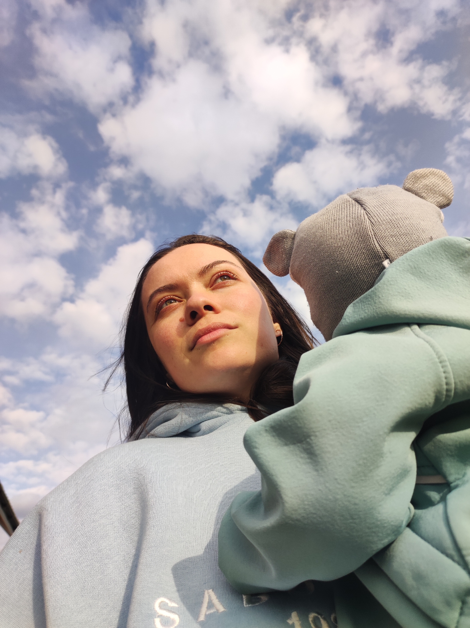 A low-angle shot captures a young woman and a child or infant against a bright, partly cloudy blue sky, likely in Фоки, Russia. The woman, with long dark hair, occupies the left side of the frame, looking upwards and slightly to her right. Her face is illuminated by warm, directional sunlight, highlighting her features and creating shadows on her right cheek. She has light-colored eyes and a mole visible on her left cheek.
She is wearing a light blue hooded sweatshirt, with partial text visible on the front that appears to say "SA" and "10". Her left ear is visible, adorned with a small hoop earring.
To the right, partially obscuring the woman's right shoulder, is the back of a child's head, wearing a grey knitted hat with small ear-like protrusions. The child, or a baby being carried, is dressed in a slightly darker, more teal-colored hooded sweatshirt. The perspective suggests the child is being held or worn in a carrier by the woman, looking in the same general direction as her.
The background is dominated by a vast blue sky with numerous scattered white cumulus clouds, indicating daytime, possibly late morning or afternoon given the strong sunlight. The overall mood of the image is serene and contemplative, with the subjects gazing upwards towards the sky.