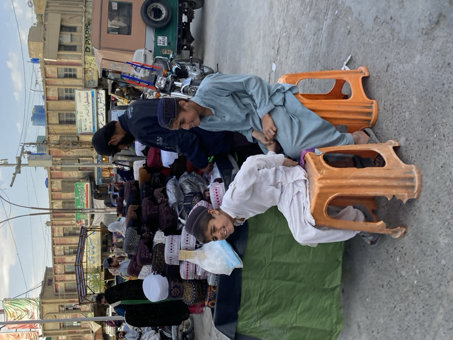 This image captures a vibrant street market scene in Quetta City Tehsil, Pakistan. In the foreground, two young boys are seated on orange plastic stools. The boy on the left wears a white shalwar kameez and a dark cap, smiling broadly and looking towards the right. His stool exhibits damage to its lower base. The boy on the right wears a light blue shalwar kameez and a dark cap, also looking right with a slight smile. Directly behind them, a market stall, covered by a green tarp, displays a large assortment of traditional embroidered caps (topis) in various colors and designs, some packaged in clear plastic. An adult male, wearing a dark shirt and cap, stands behind the stall, facing left, appearing to attend to the merchandise. To his left, another individual is partially visible.
In the midground, a green utility vehicle, possibly a rickshaw, is parked, featuring a spare tire mounted on the rear and a license plate partially visible as "9514." Adjacent to it is a motorcycle with "Traffic Police" decals on its front fairing. The background reveals multi-story urban buildings, appearing somewhat aged, housing shops and residences. Visible signs on these buildings include "NATIONAL" and "D PAPIS," along with others in Urdu script. Several blue water tanks are visible on the building rooftops. Overhead, utility poles and electrical wires crisscross against a light blue sky with scattered clouds. The ground is paved, consistent with an urban street or marketplace. Numerous other pedestrians are visible in the background, moving through the market environment.