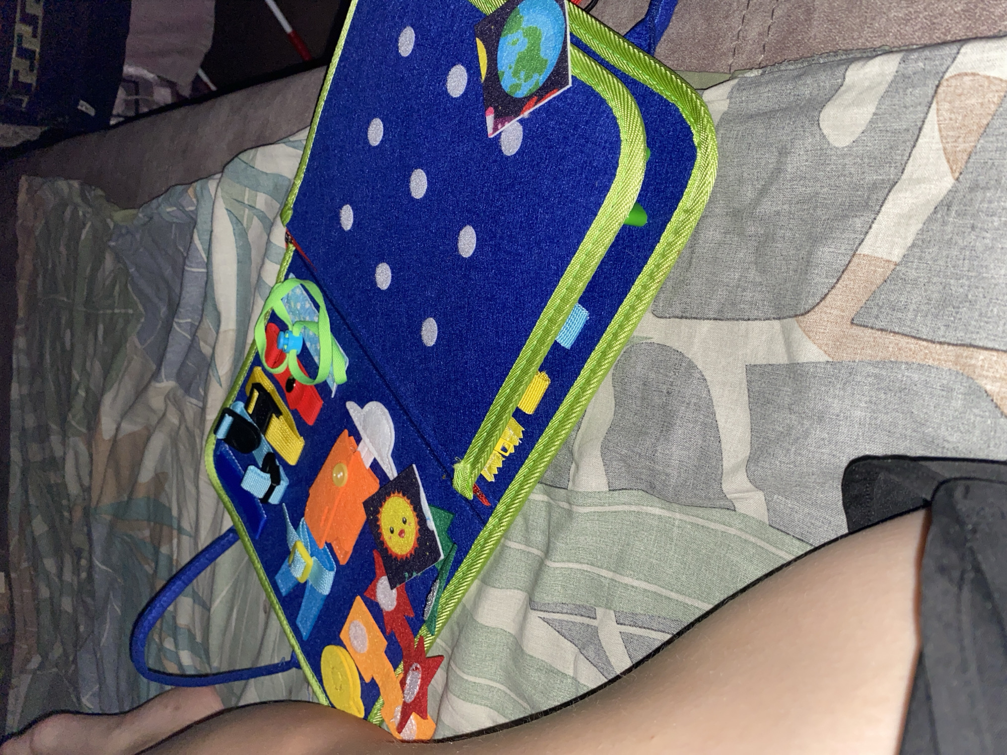An open, blue felt busy board lies on a bed with a patterned duvet. The busy board is adorned with various small, colorful felt appliques and interactive elements, including shapes, buckles, and zippers. A blue fabric handle is attached to one side of the board. A person's bare leg and a portion of black shorts are visible in the lower left corner of the frame, resting on the bedding. The background consists of draped bedding in muted tones. The lighting suggests it is nighttime or a dimly lit room. The scene is indoors and does not contain any elements that would indicate a specific location in the city of Nikolo-Pavlovskoye, Russia.