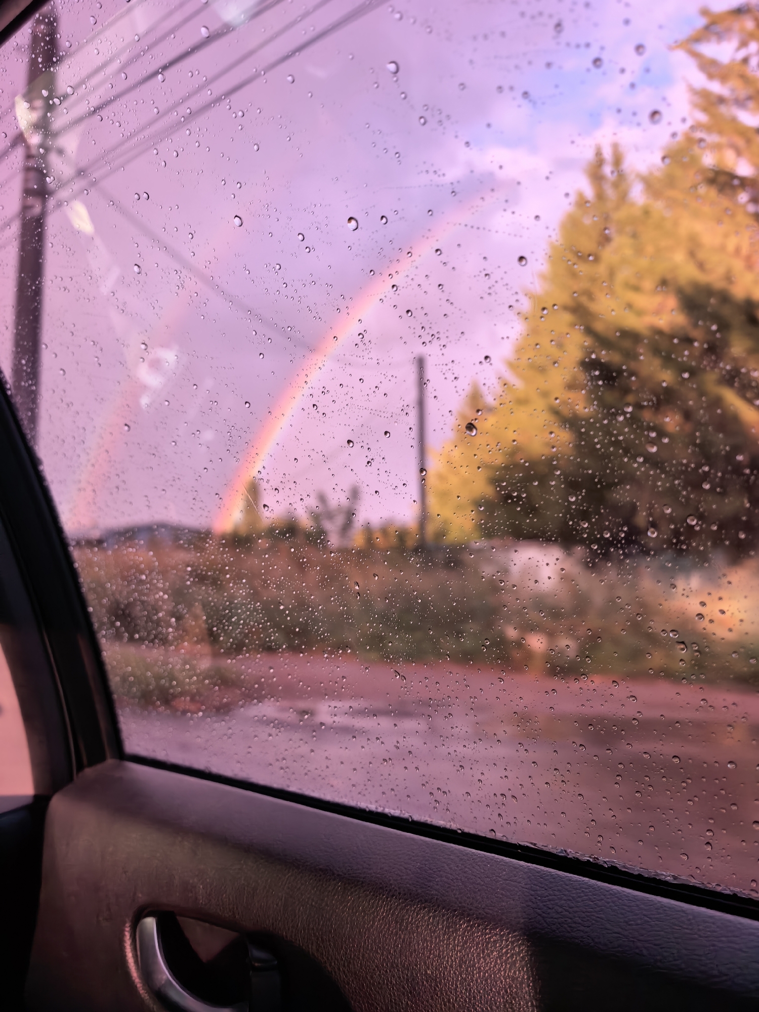 The image shows a view through a car window on a rainy day. The window is covered in water droplets, distorting the view of the outside. Through the rain-streaked glass, a rainbow is visible against a pinkish-purple sky. In the background, there are trees with yellow-green foliage and a hint of a rural landscape. The interior of the car, specifically the door panel and handle, is visible in the foreground, indicating the perspective is from inside the vehicle. The overall mood is atmospheric and somewhat melancholic due to the rain and the soft, diffused light. The image does not provide any specific information about the city of Sterlitamak, Russia.