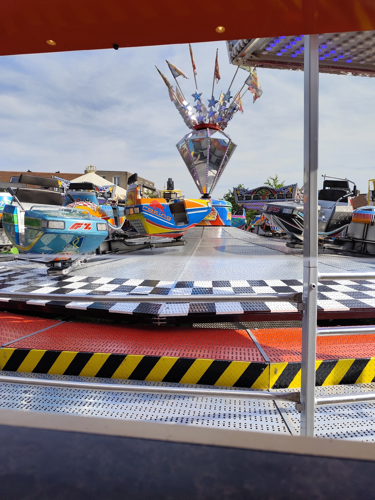 The image displays a section of a fairground ride, likely a spinning amusement ride with multiple car-like pods designed to resemble race cars. The pods are brightly colored in blue, orange, and yellow, with some featuring logos like "F1" and "Red Bull." The ride platform is covered in a checkered black and white pattern. The ride is situated outdoors, with a partly cloudy blue sky visible in the background. In the distance, buildings with tiled roofs, characteristic of residential areas in the Netherlands, can be seen. The overall impression is that of a typical funfair attraction. The location is identified as Dongen, Netherlands, though there is no specific landmark or context in the image to confirm this definitively. There are no people visible in the image, and the ride appears to be stationary. The image is taken from a low-angle perspective, focusing on the ride's structure and decorative elements.