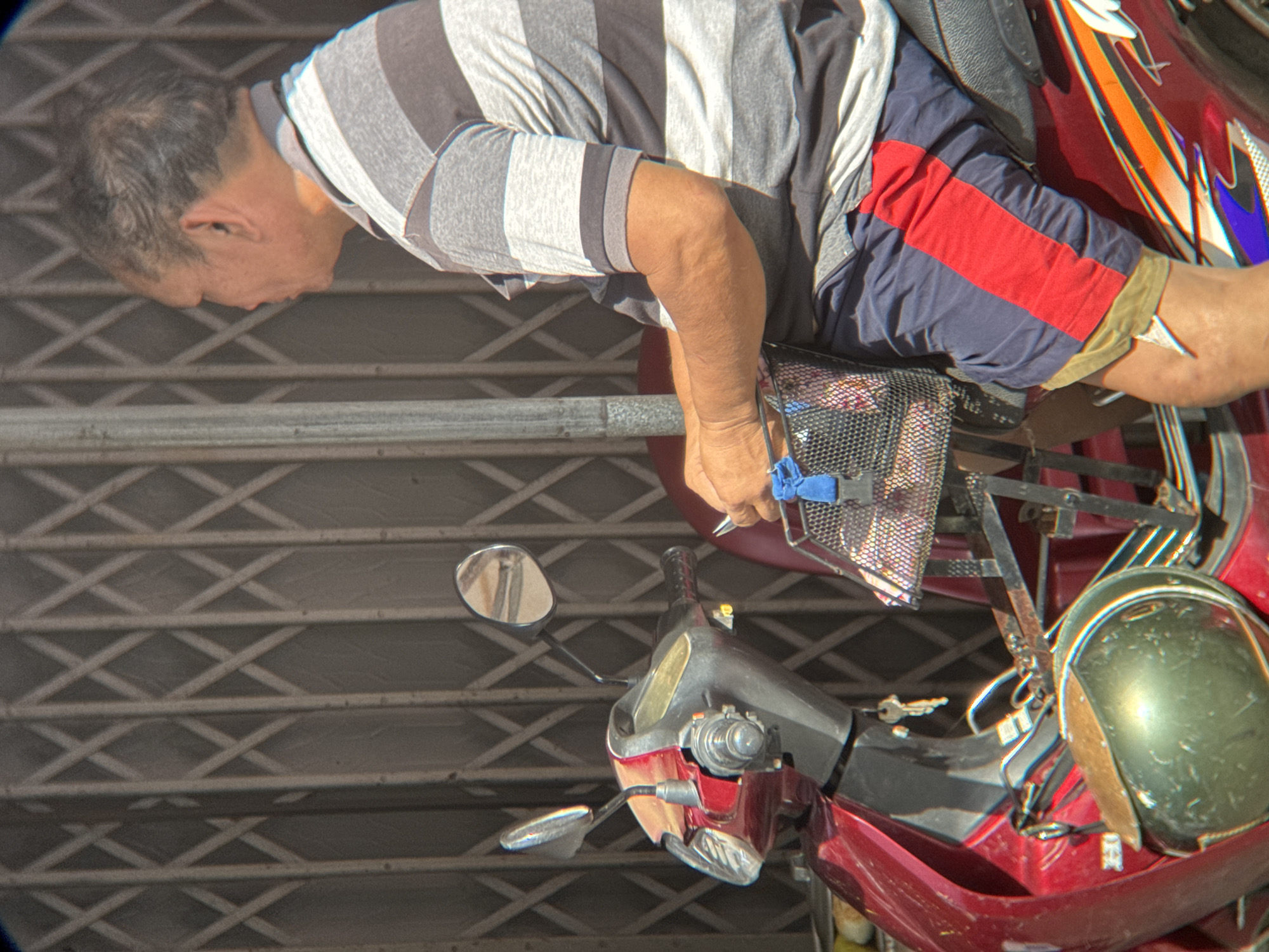A middle-aged East Asian man with thinning grey hair is seated on a red and silver motorcycle, facing right. He is wearing a grey and white horizontally striped t-shirt and navy blue shorts with a red stripe down the side. His right arm is resting on a black basket attached to the front of the motorcycle, which contains various items, including a pair of scissors and a blue ribbon. The motorcycle has a silver mirror on the right handlebar and a red body with decorative orange and purple graphics. A key is hanging from the ignition. Behind the motorcycle is a grey, collapsible metal gate. The scene appears to be an outdoor setting, possibly a street or alleyway, in Nha Trang, Vietnam, suggested by the presence of a motorcycle, the typical architecture of a metal gate often seen in Vietnamese cities, and the clothing of the individual. The lighting indicates it is daytime.