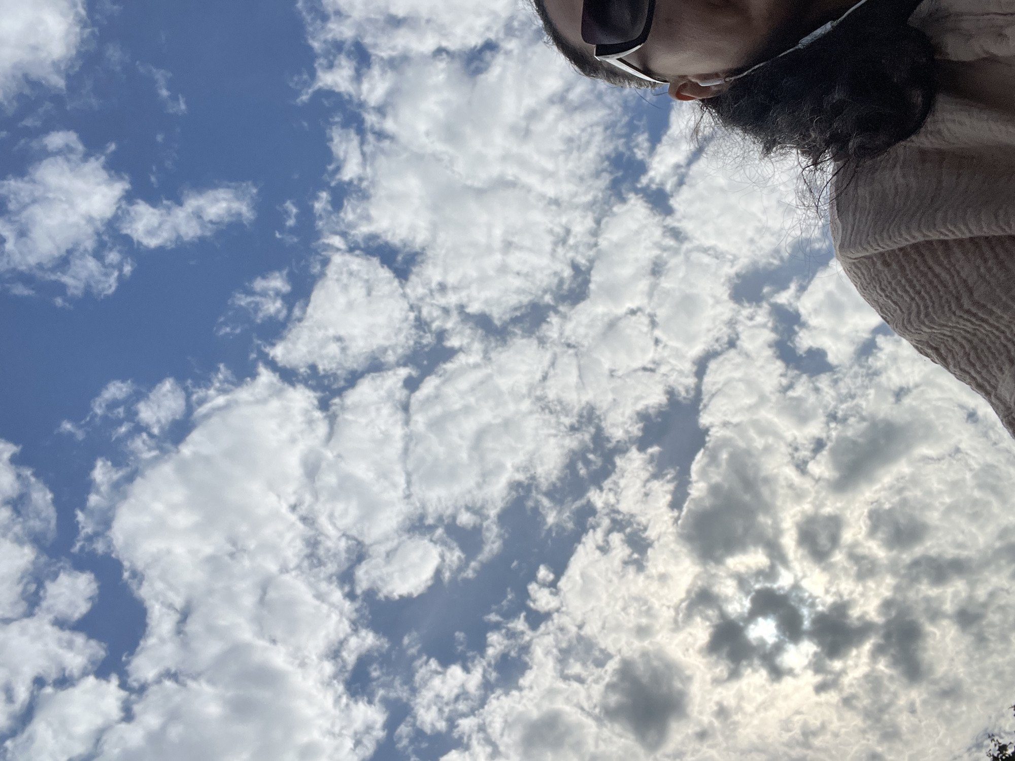 This image captures a low-angle view of a partially overcast sky over Tarnivtsi, Ukraine. The sky features a vibrant blue background interspersed with numerous cumulus and stratocumulus clouds, appearing in shades of white and light grey. A bright light source, consistent with the sun, is obscured by denser cloud formations in the lower-left quadrant, creating a radiant highlight. In the bottom-right section of the frame, the right side of an individual's head and upper shoulder are visible. The person has dark, curly hair and wears dark, rectangular sunglasses featuring distinctive white frames. A white wired earbud is inserted into their right ear. They are clad in a light, possibly beige or pinkish-tan, textured garment, resembling a knit sweater or shawl, draped over their right shoulder.