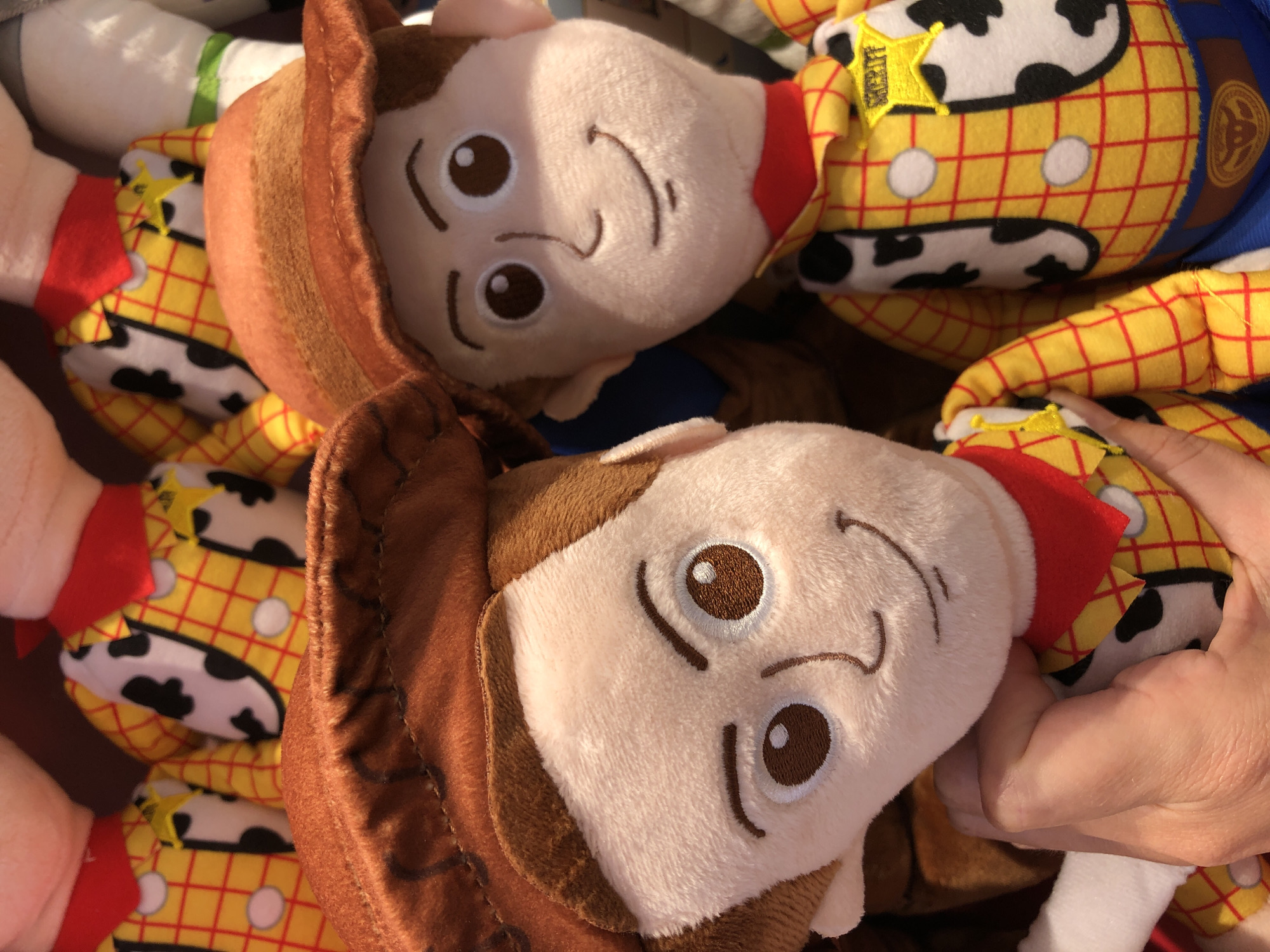 The image features a close-up view of several plush toys resembling Woody from Toy Story. The toys are densely packed together, with some partially obscuring others. The primary subject is the faces and upper bodies of the Woody dolls, characterized by their signature yellow and red plaid shirts, brown cowboy hats, and embroidered facial features.
In the foreground, a doll's face is prominently displayed, with large, round brown eyes and a smiling mouth. Its brown hat is tilted, revealing part of its plush head. To the right, another Woody doll is partially visible, held by a hand, suggesting someone is interacting with the toys. This hand is fair-skinned and shows some detail in its fingers.
In the background and interspersed between the main subjects, other Woody dolls are visible. Their distinctive sheriff's badges are noticeable on some of their vests. The texture of the plush material is evident, with a soft, fuzzy appearance. The overall color palette is dominated by the warm tones of brown, yellow, and red, consistent with Woody's attire.
The lighting appears to be natural, suggesting an indoor setting with ambient light, possibly a store or a home environment where these toys are displayed or stored. There is no indication of specific weather conditions or time of day, though the brightness suggests daytime. The context provided of "Đà Nẵng, Vietnam" places the scene geographically. No discernible text is present within the image itself, beyond the embroidered details on the toys. The arrangement of the toys and the presence of a hand imply a casual handling or display of the merchandise.